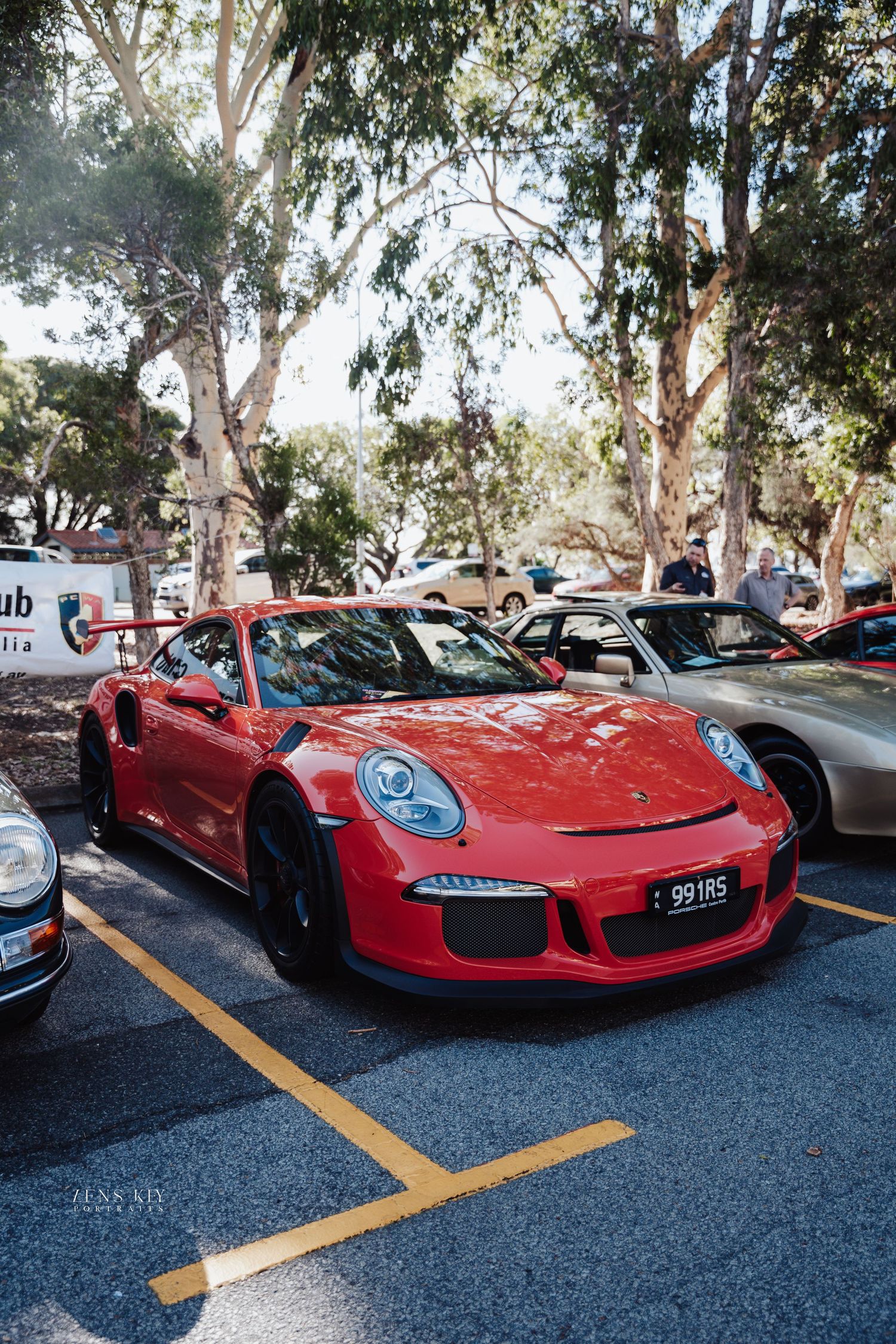 Red Porsche 911 GT3 RS sports car parked in a shaded lot at an outdoor car show.