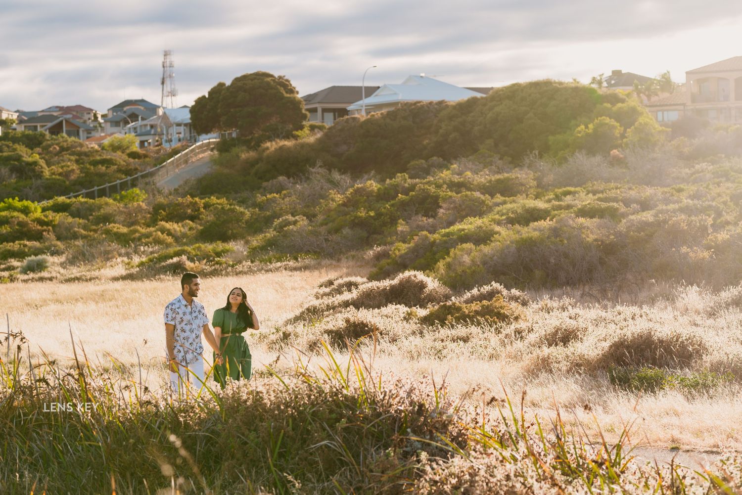 Couple walking through golden coastal dunes at sunset during a relaxed outdoor engagement photo session.