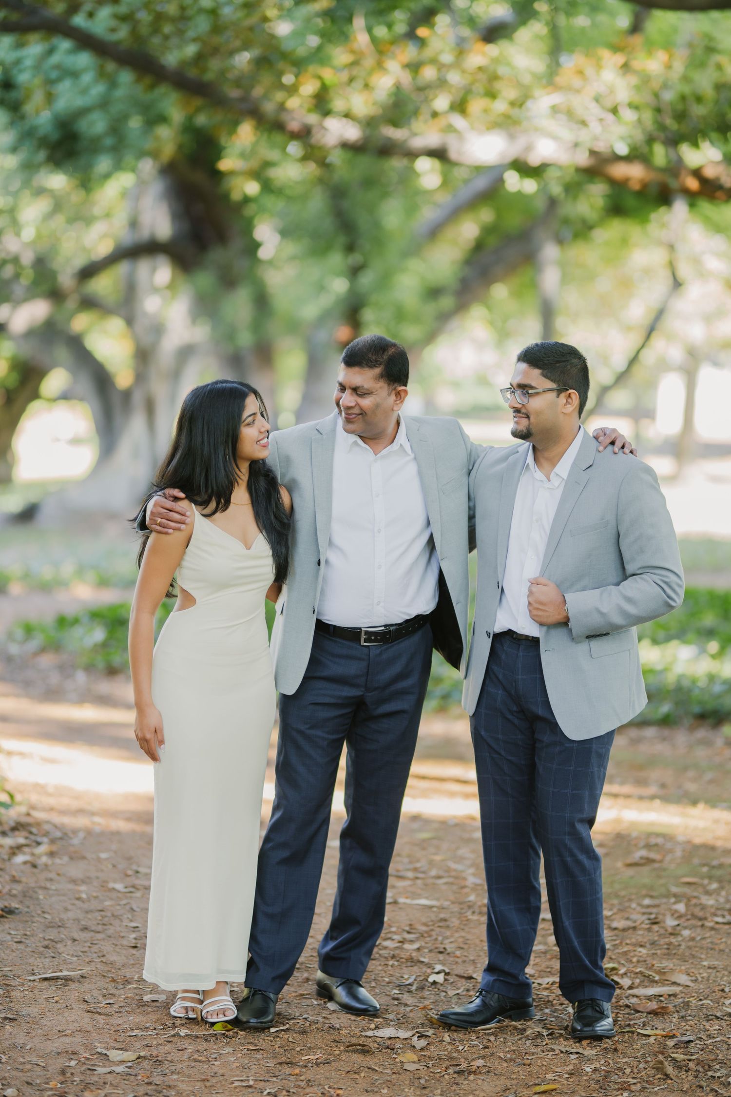 Three adults pose together smiling in a sun-dappled park with large trees in background.