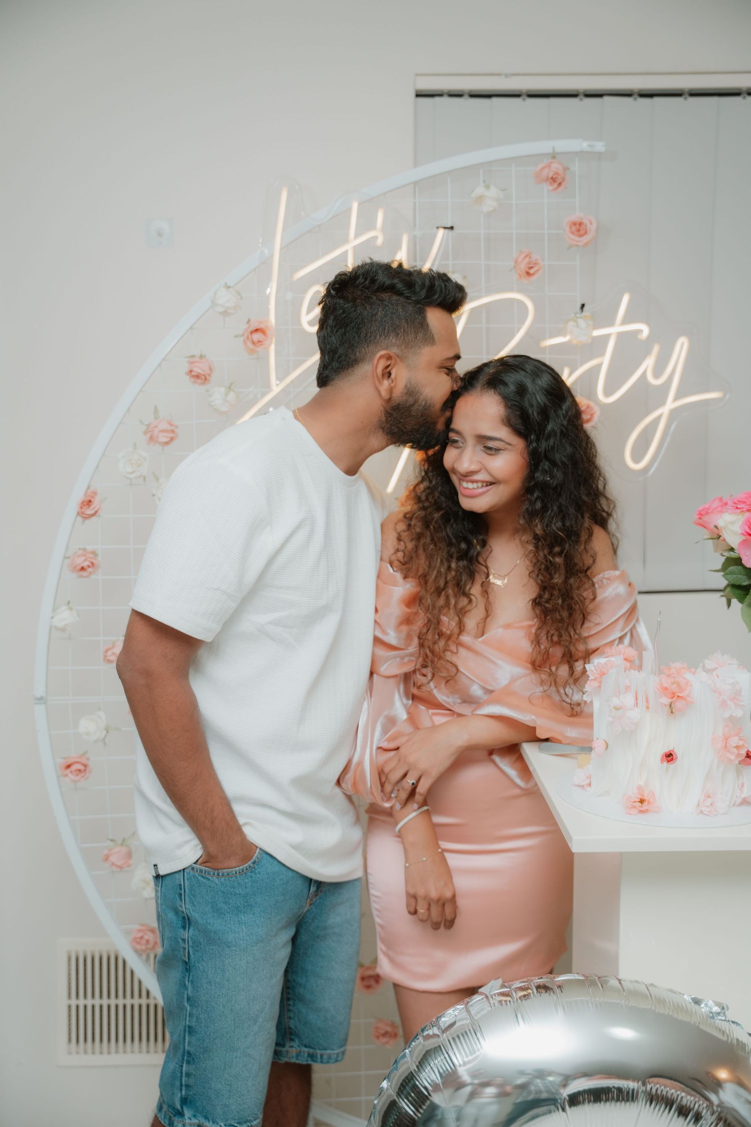 Man kissing woman on cheek at a romantic birthday party with floral Let's Party neon sign backdrop.