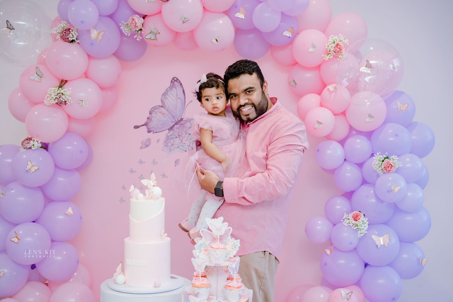 Smiling father holding baby girl in pink dress at butterfly-themed first birthday party with balloon arch.
