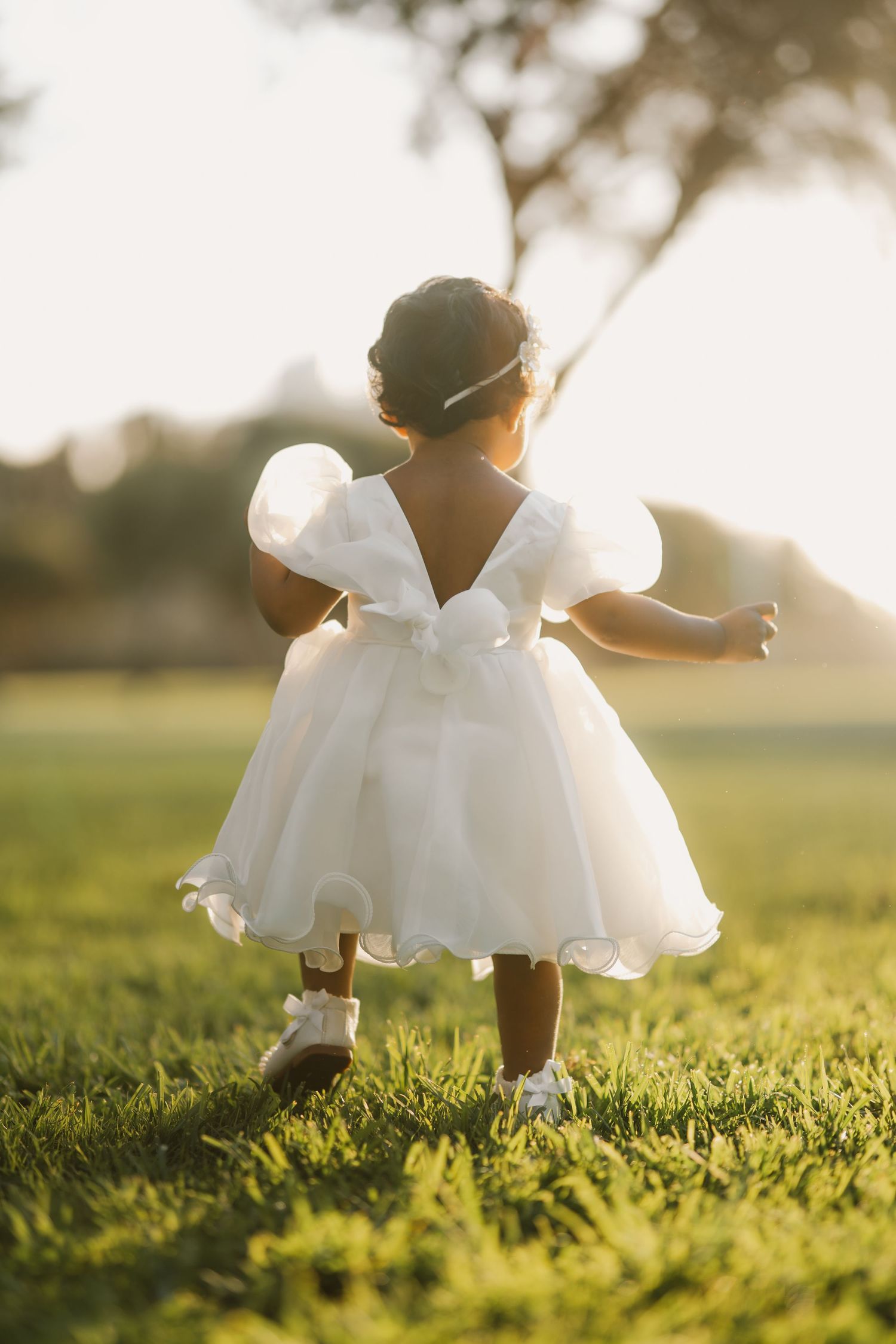 Toddler girl in white dress with bow walking away in golden sunset light.