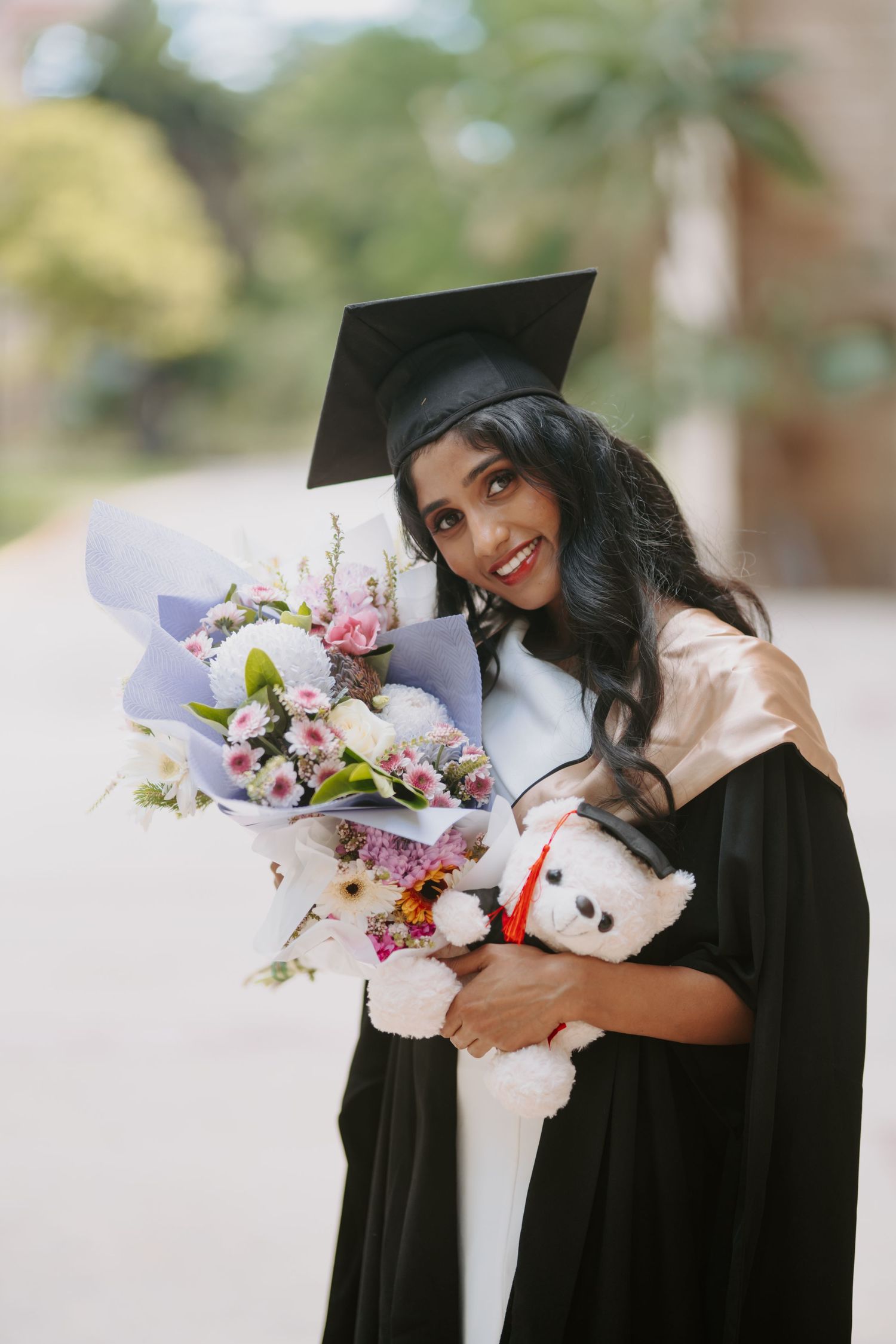 Smiling female graduate in black cap and gown holding a bouquet of flowers and a teddy bear outdoors.