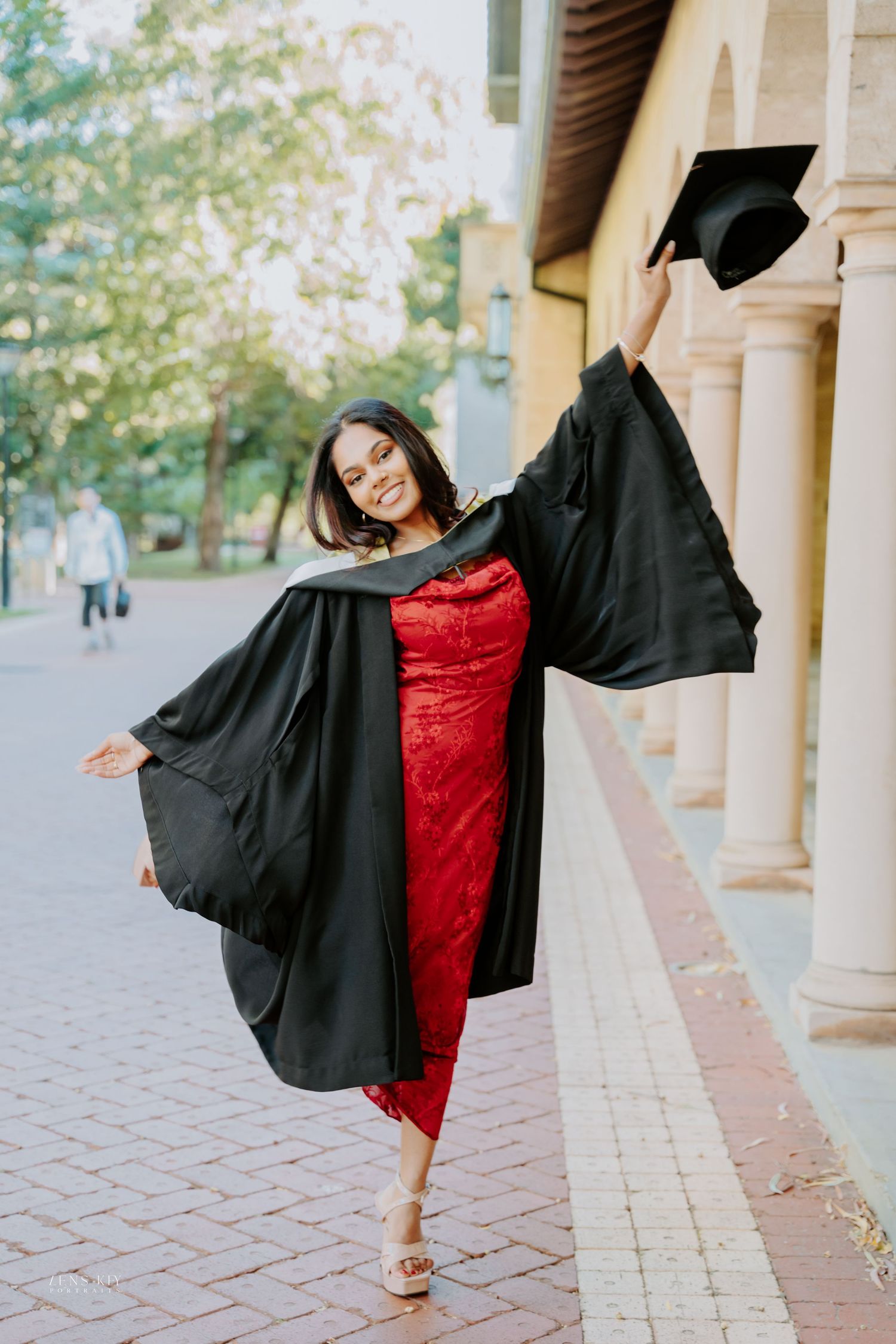 Joyful graduate in red dress and black gown raises mortarboard cap while posing outdoors.