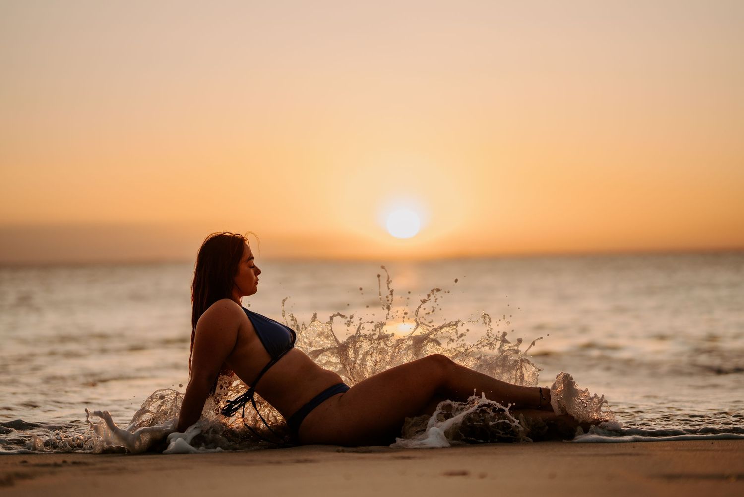 A woman in a dark bikini sits on the beach as ocean waves splash around her at sunset.