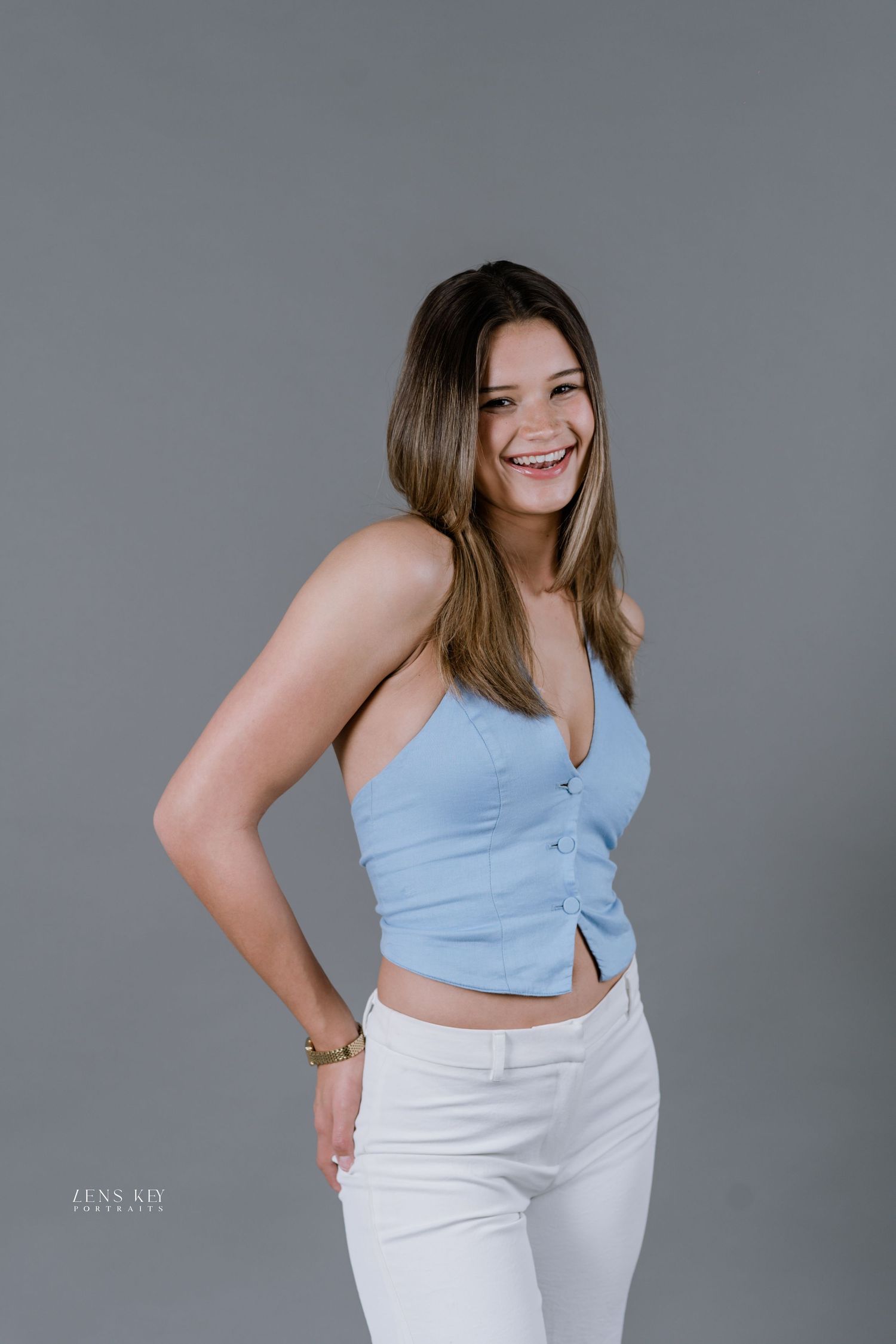 Young woman smiling in blue crop top and white pants poses confidently against gray background.