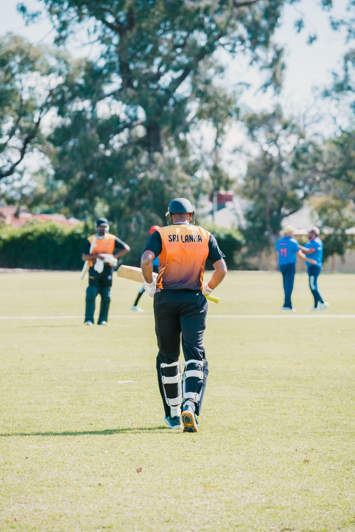 Cricket batsman in orange Sri Lanka jersey walking onto a green field wearing protective gear.