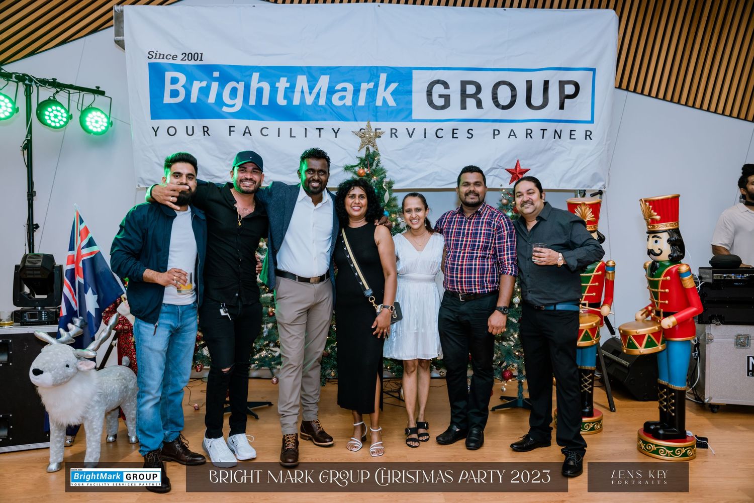 Group of seven people posing in front of BrightMark Group Christmas Party 2023 banner with holiday decorations.