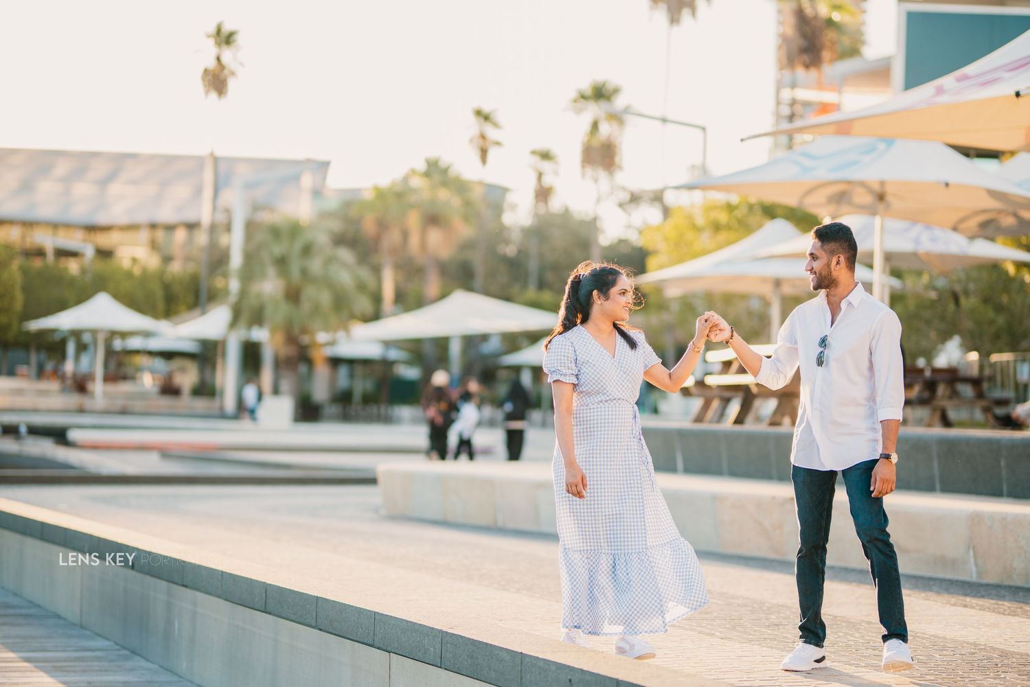 Couple playfully dancing and holding hands on a sunny urban plaza during a casual engagement photo shoot.