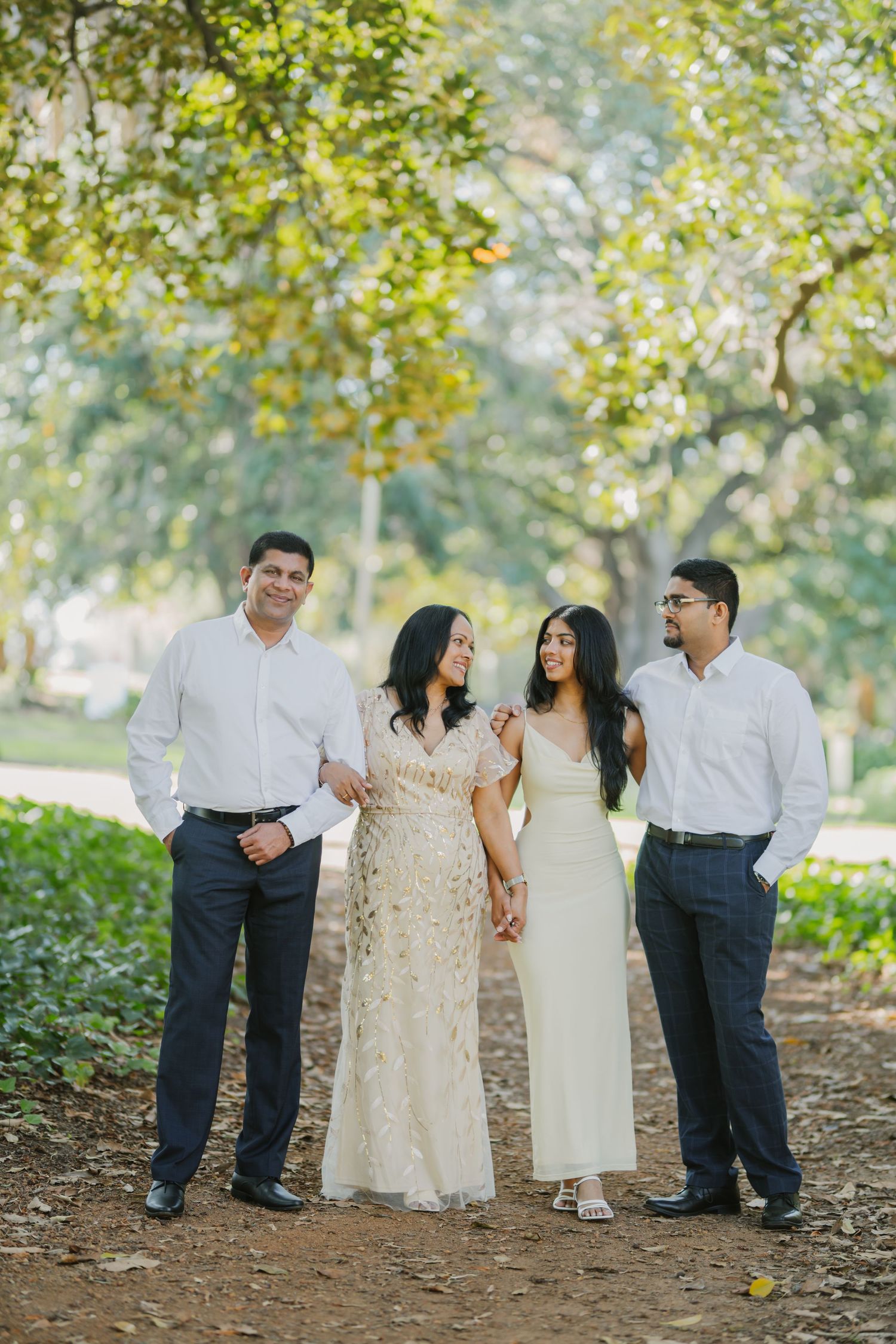 Family of four poses on a tree-lined dirt path, two women in white dresses flanked by men in white shirts.