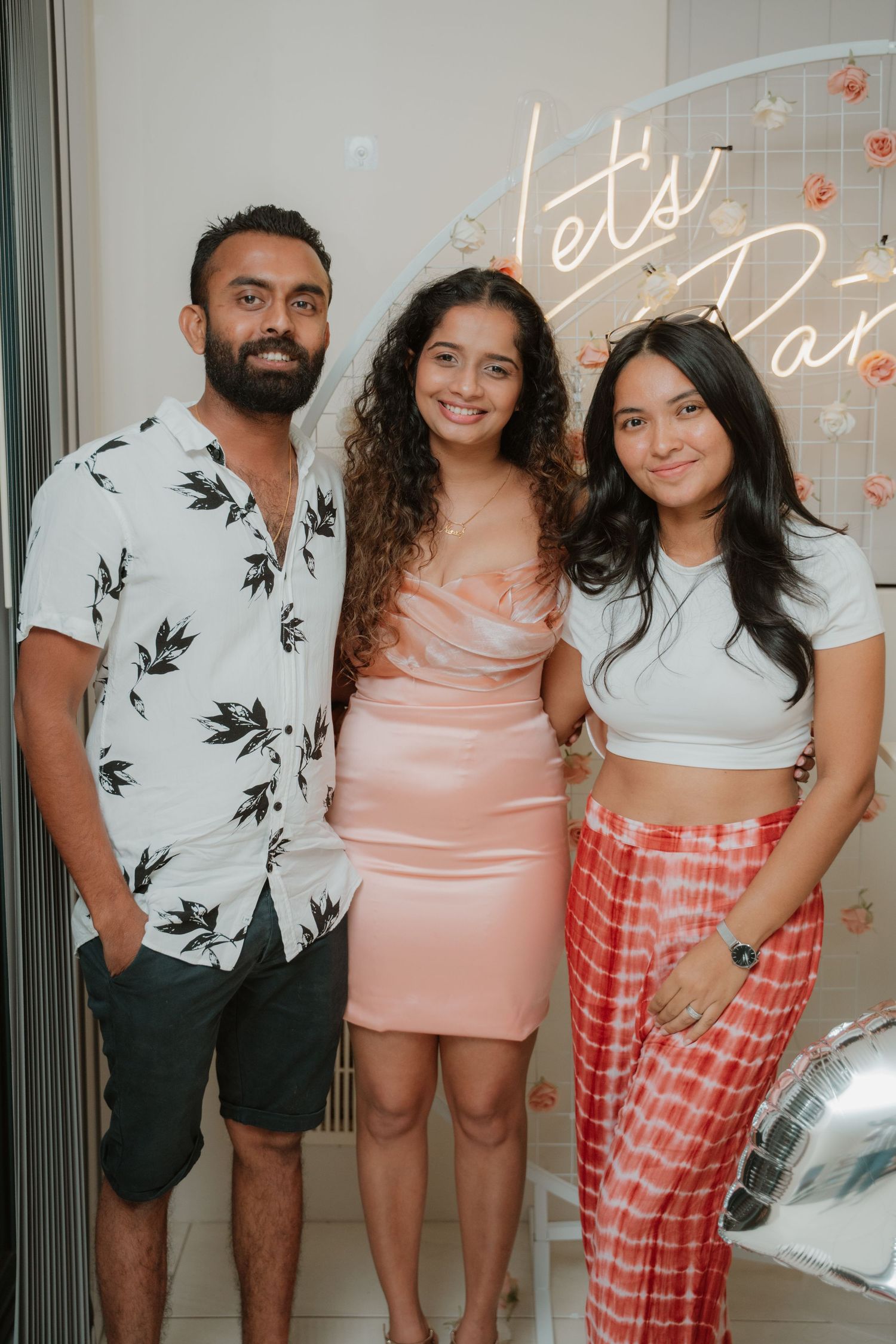 Three young adults posing together at a party with neon lets party sign.