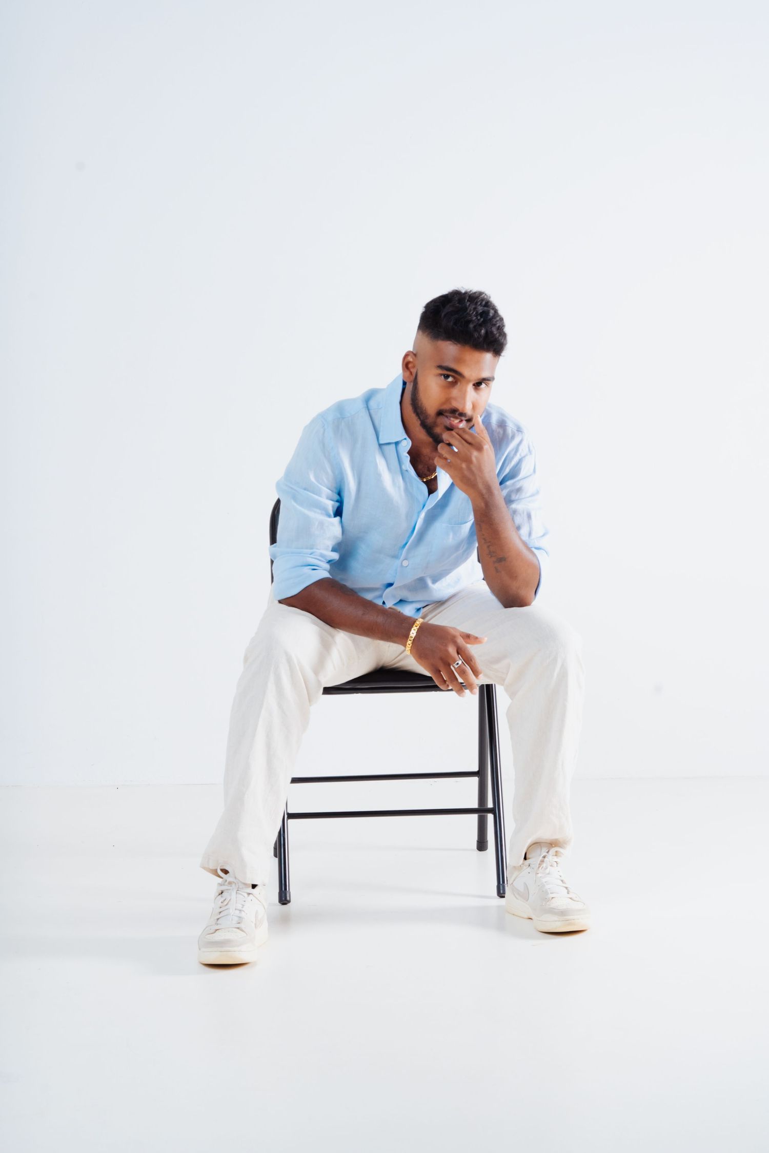 A stylish man in a light blue shirt and white pants sits on a chair with hand on chin in a white studio.
