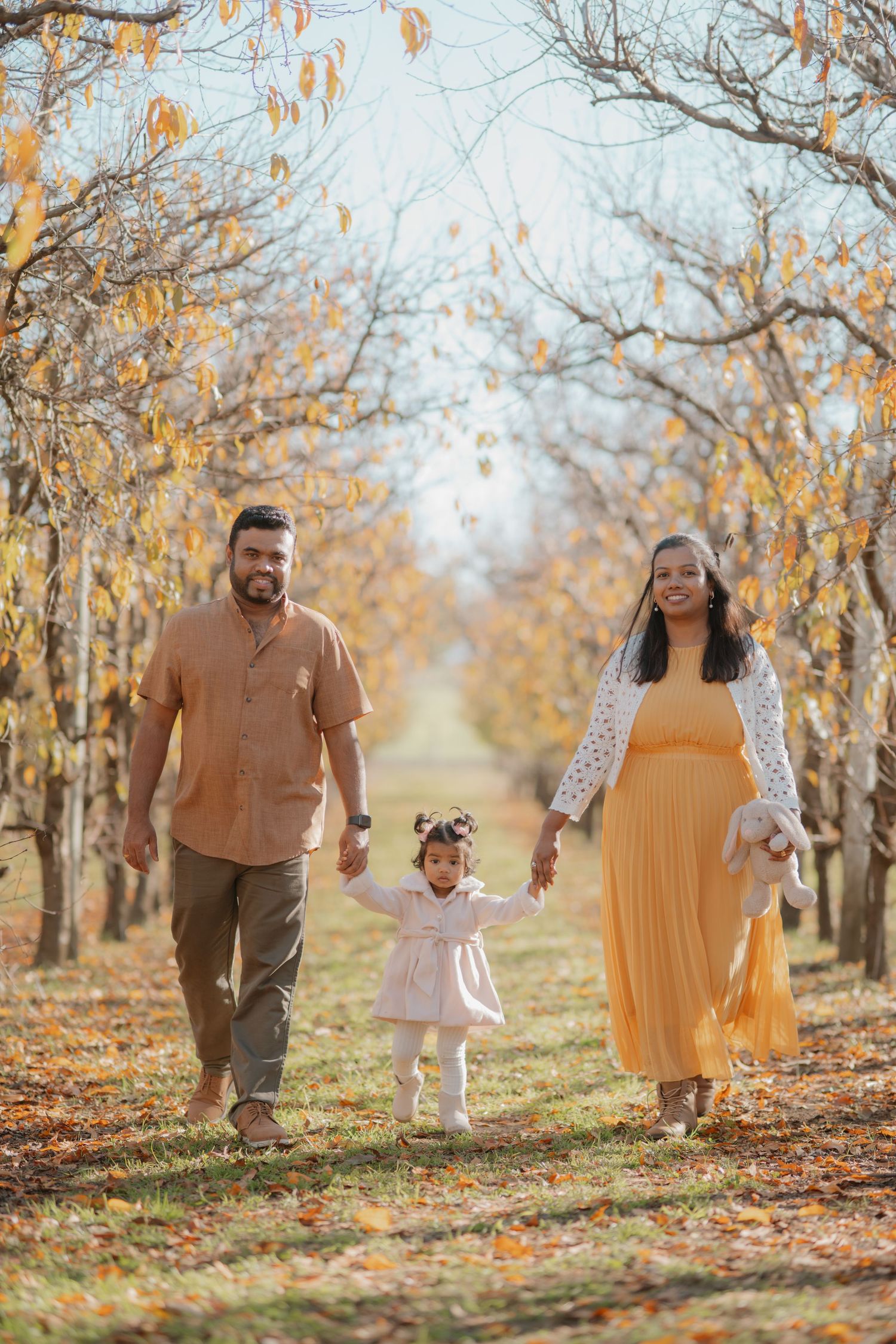 Pregnant mother in yellow dress and father hold toddler daughter's hands in autumn orchard.