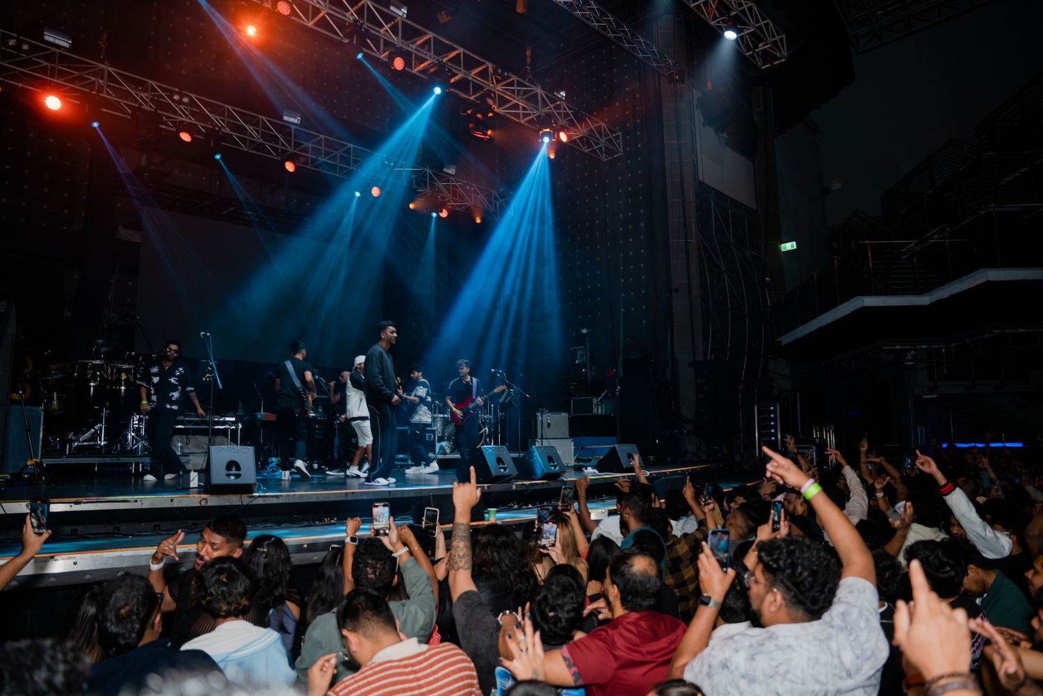 Large crowd cheering at concert with blue stage lights and performers on stage.