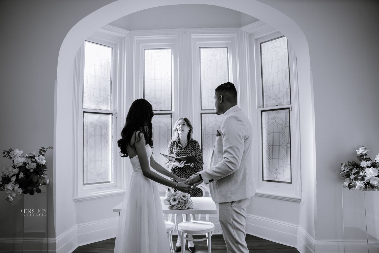 Black and white photo of couple exchanging vows with officiant by arched windows.