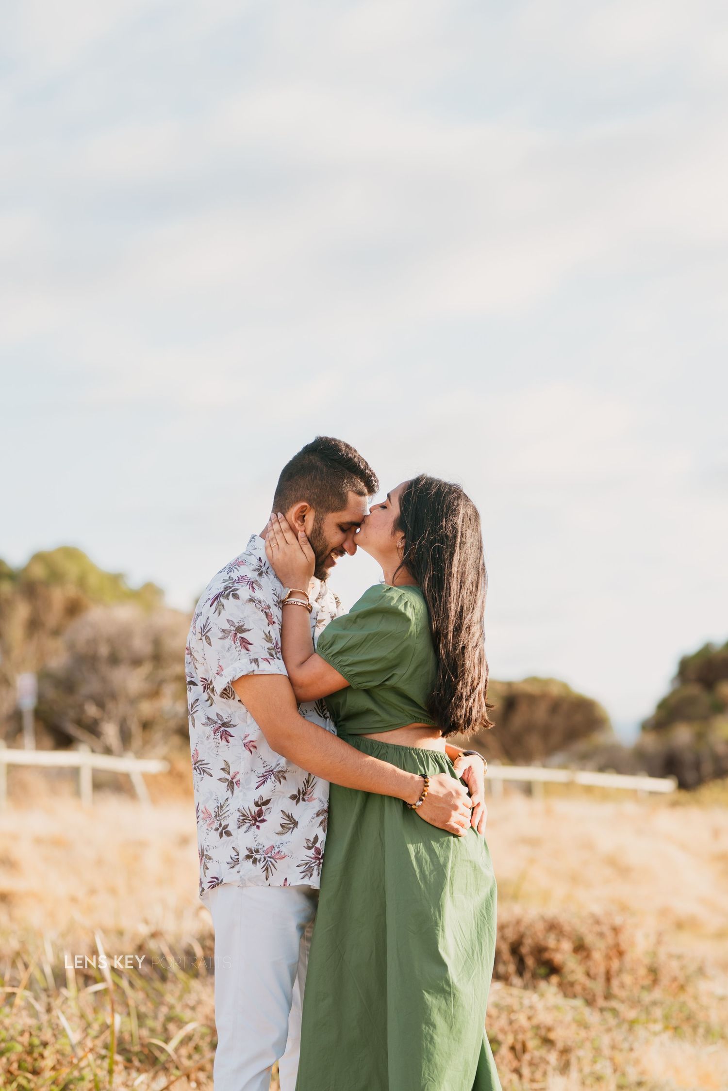 Couple sharing a romantic almost-kiss in a sunny open field, woman in green dress and man in floral shirt.