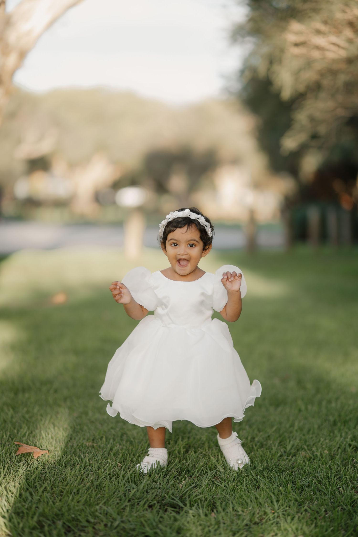 Happy toddler girl in white tulle dress standing on green grass outdoors.