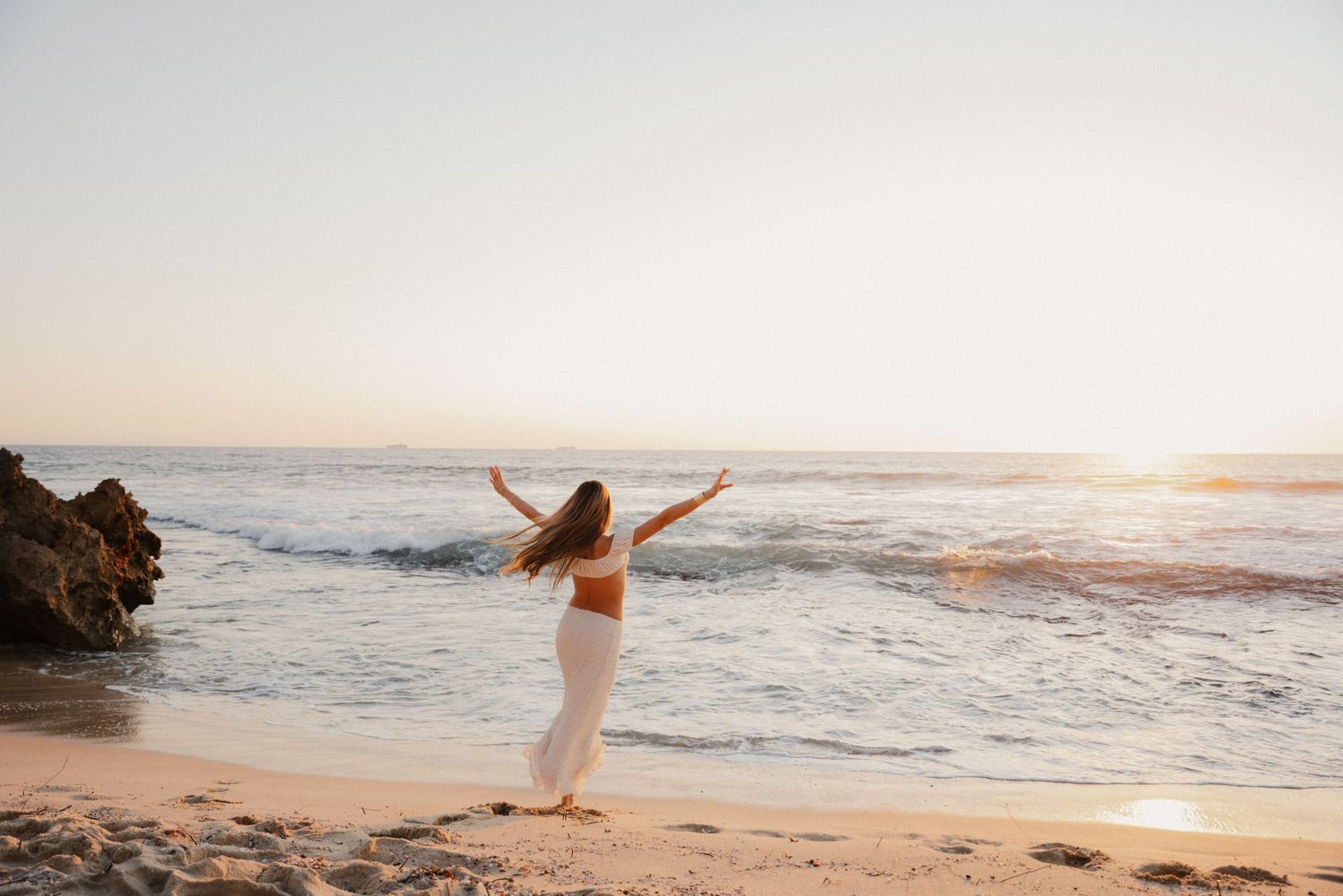 Woman in white outfit dancing freely on a sandy beach at golden sunset with ocean waves behind her.