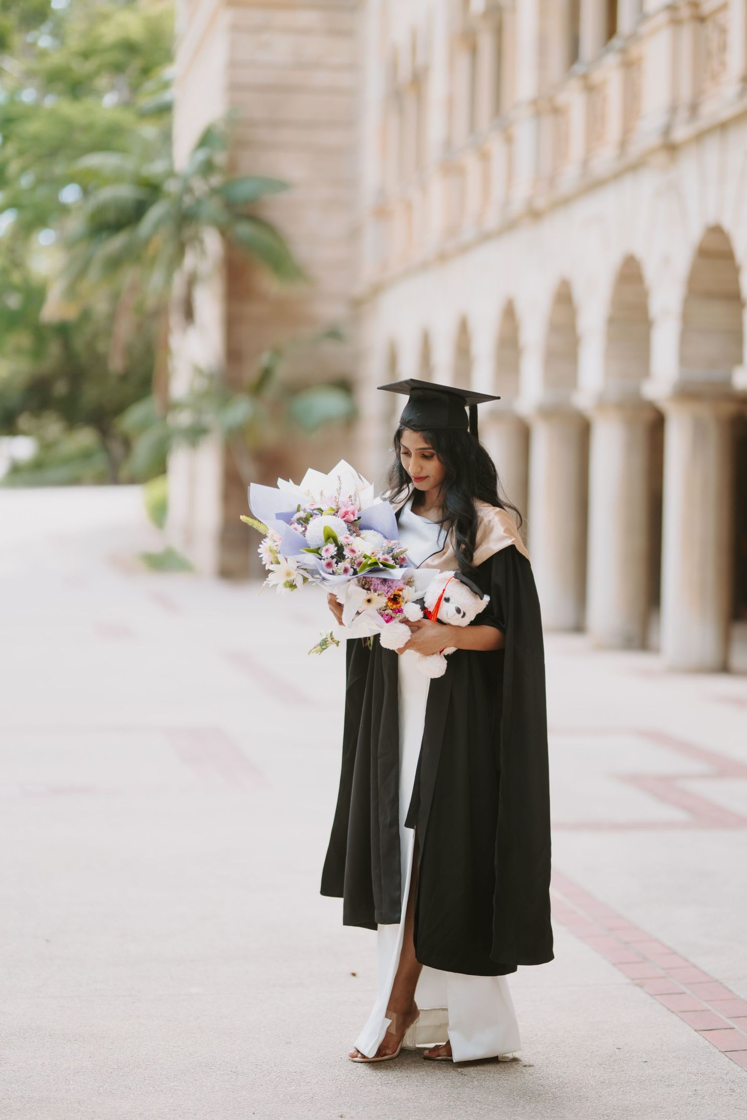 Female graduate in black cap and gown holding a large bouquet and teddy bear in a courtyard.