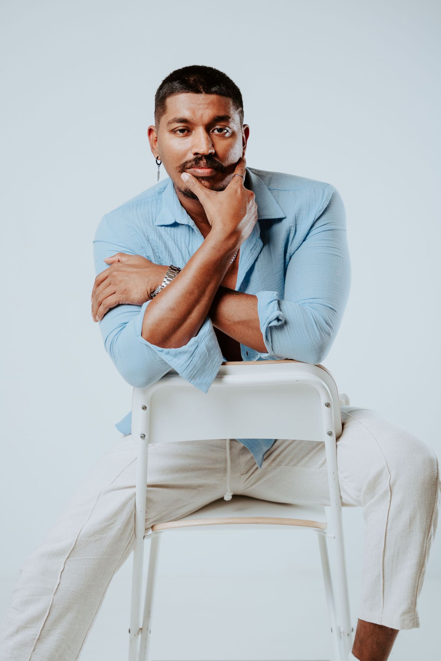 Man in light blue shirt leans thoughtfully on white chair back in bright studio setting.