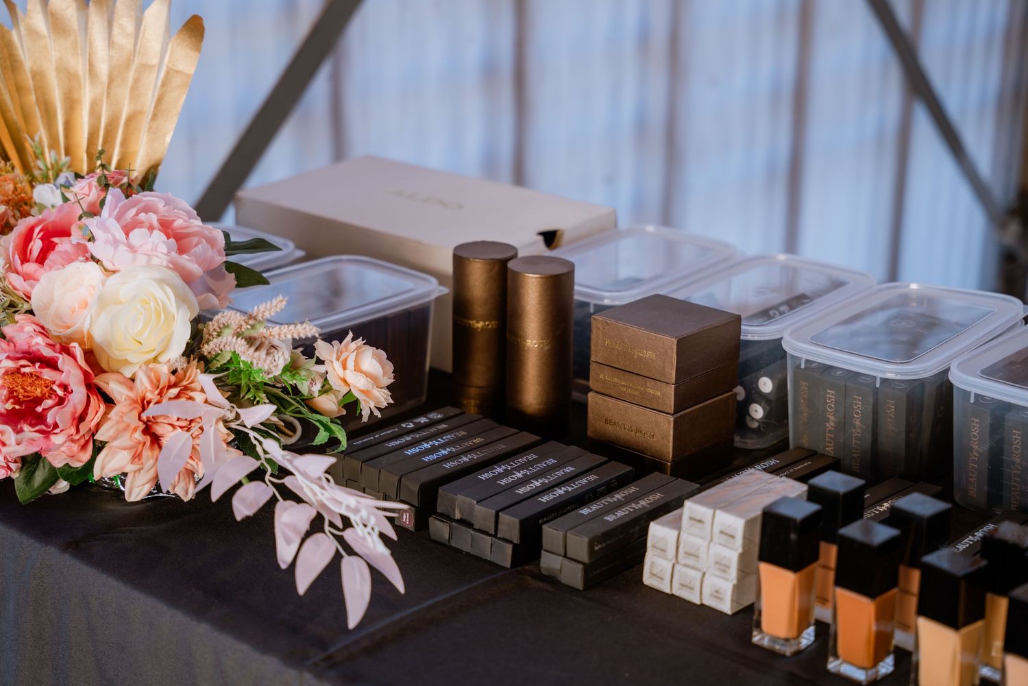 A makeup artist station with bronzers, foundations, and pink floral arrangement on a dark table.