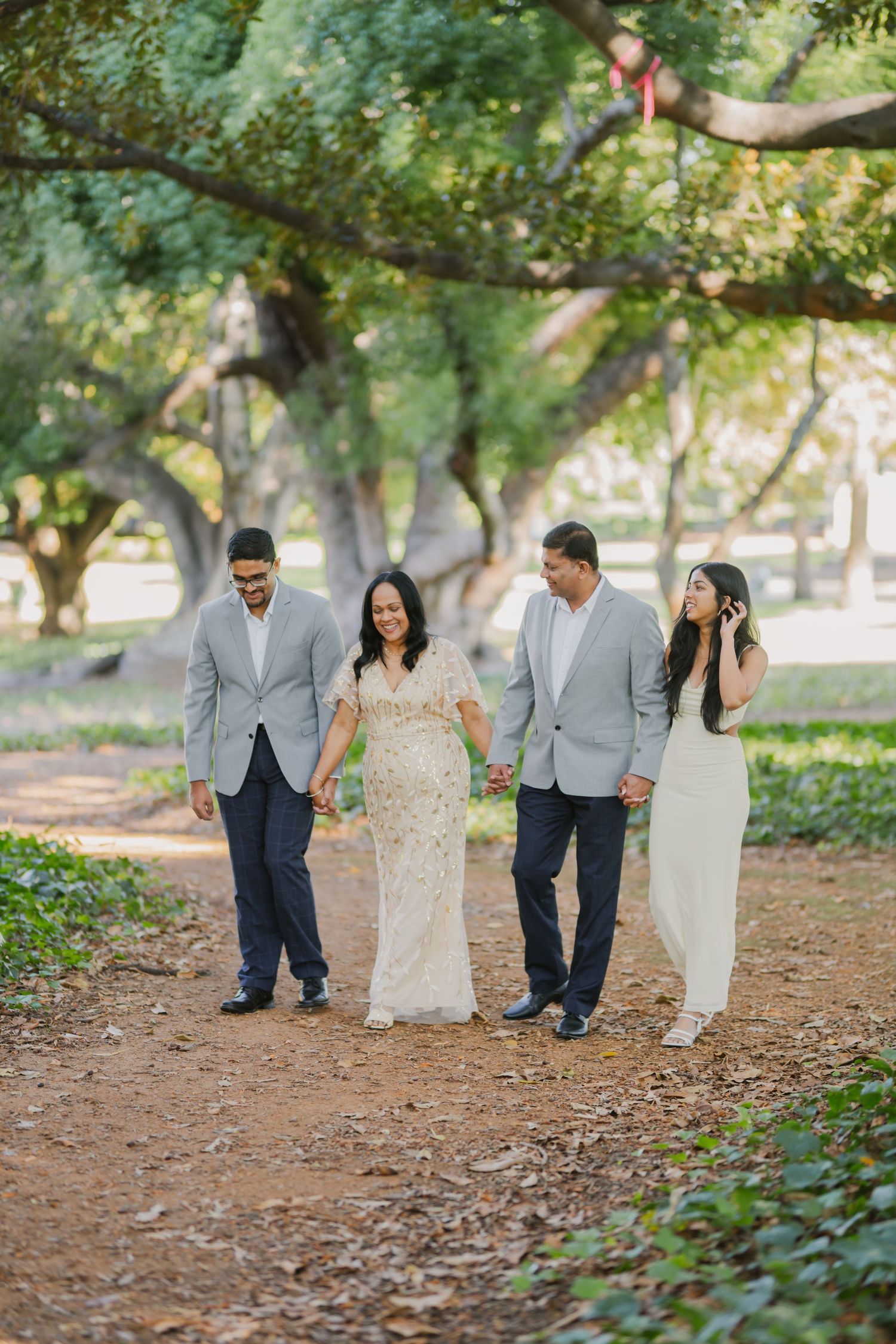 Family of four walks hand-in-hand along a shaded dirt path surrounded by large green trees.