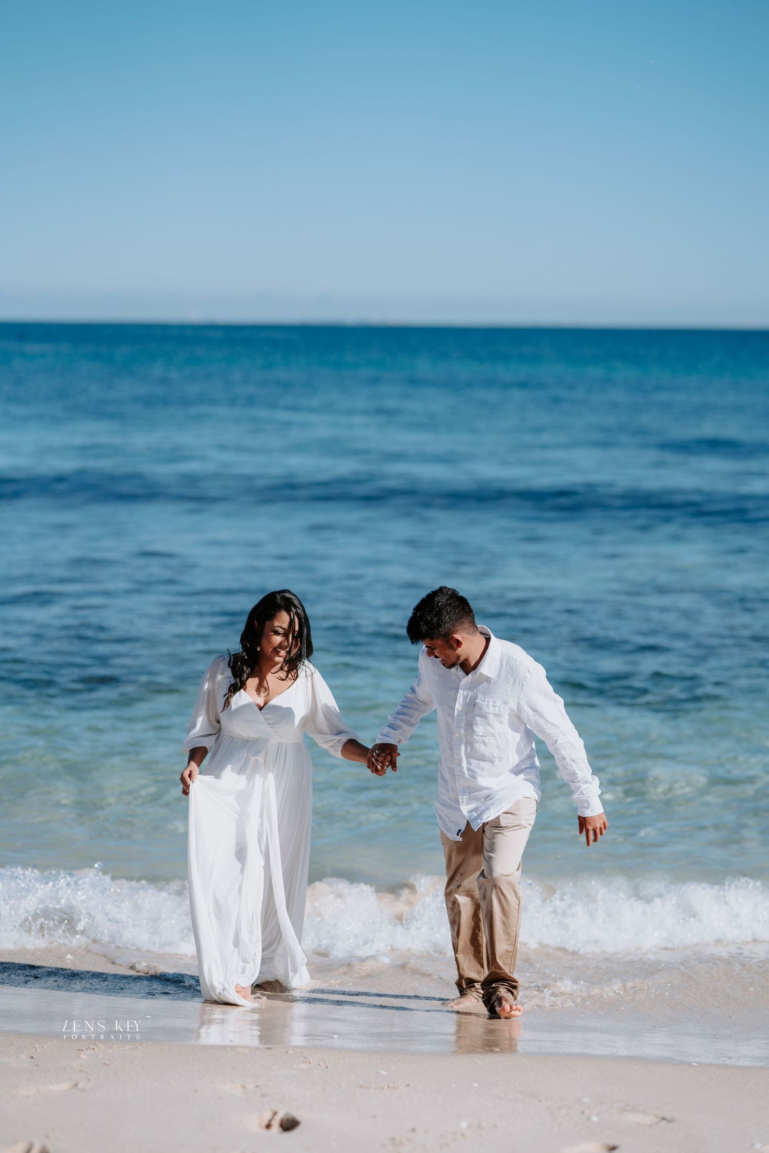 Joyful couple holding hands walking along a stunning turquoise beach shoreline in white outfits.
