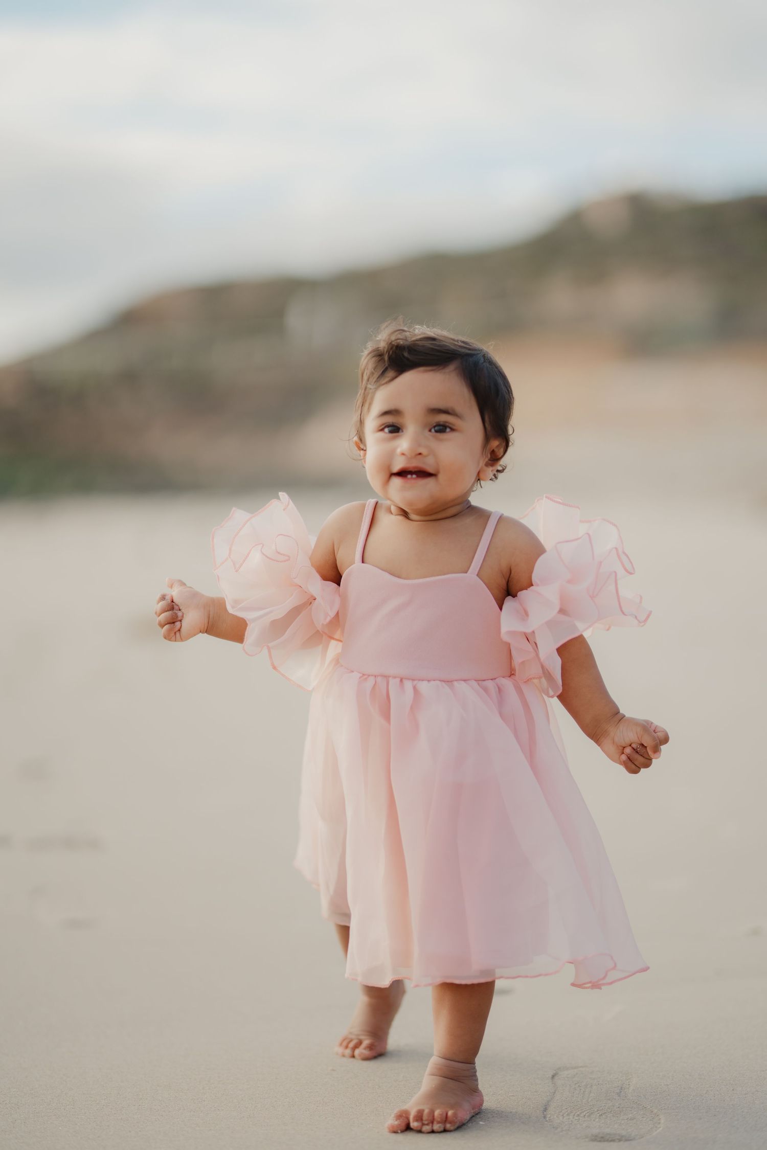 Smiling baby girl in pink ruffle dress standing barefoot on sandy beach.
