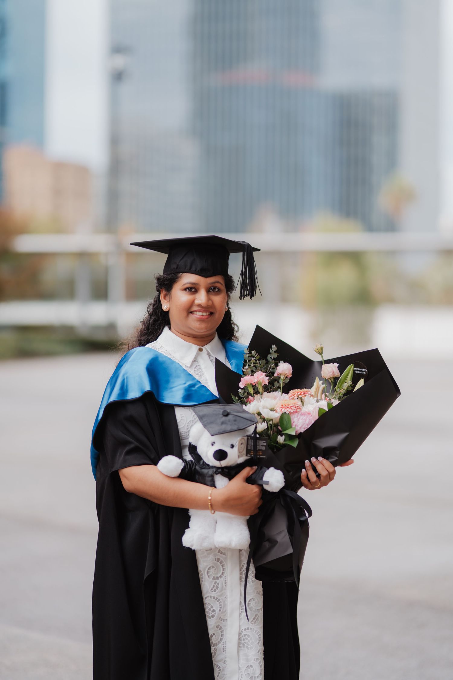 Smiling woman in black graduation gown and cap holds flowers and teddy bear outdoors.