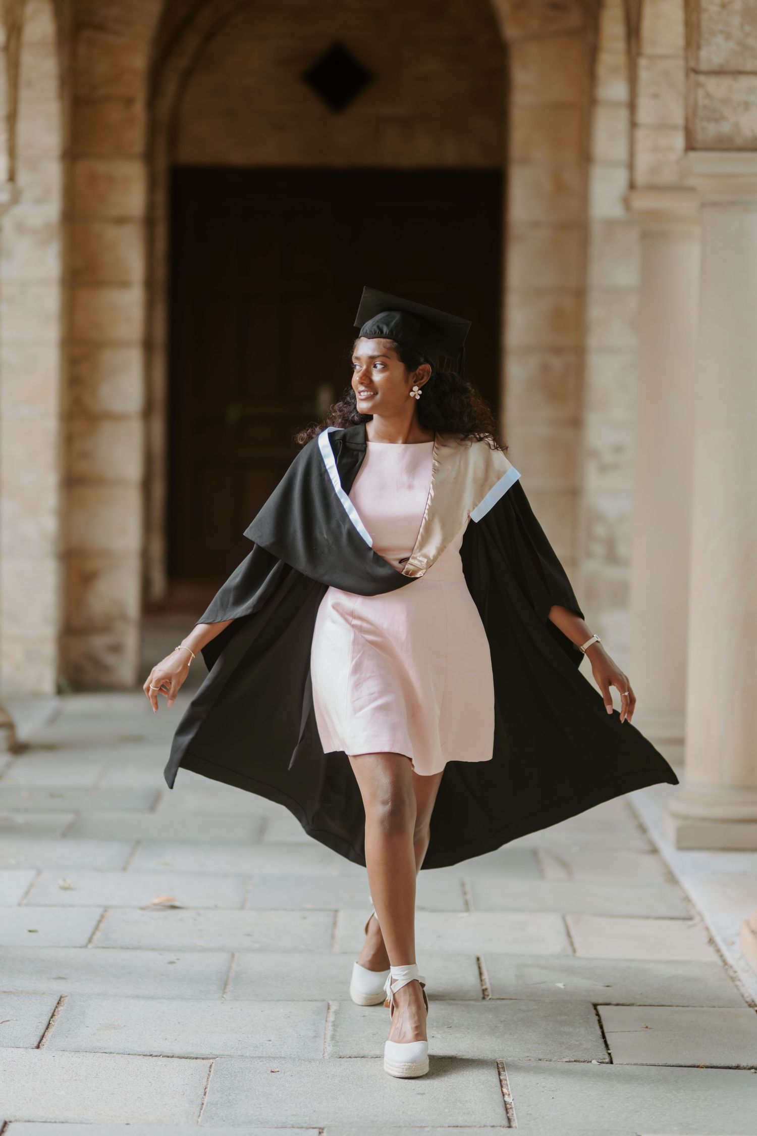 Young woman in black graduation gown and cap walks confidently through stone archway columns.
