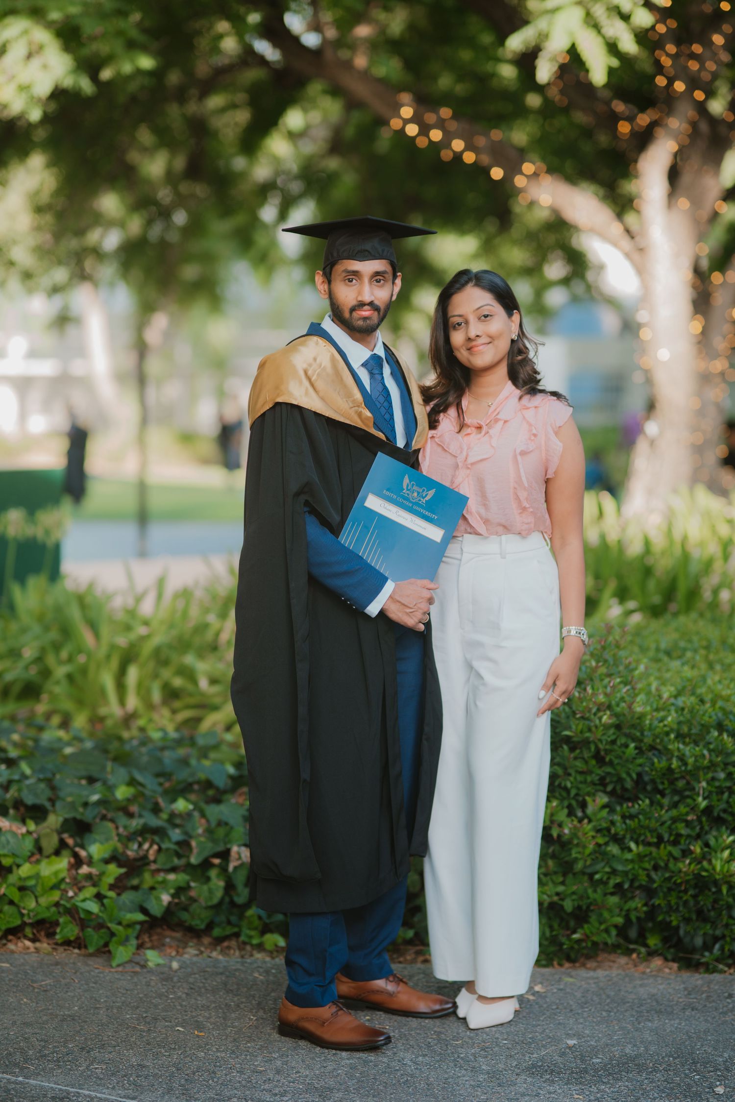 Male graduate in black and gold gown holding diploma posing with woman in park setting.