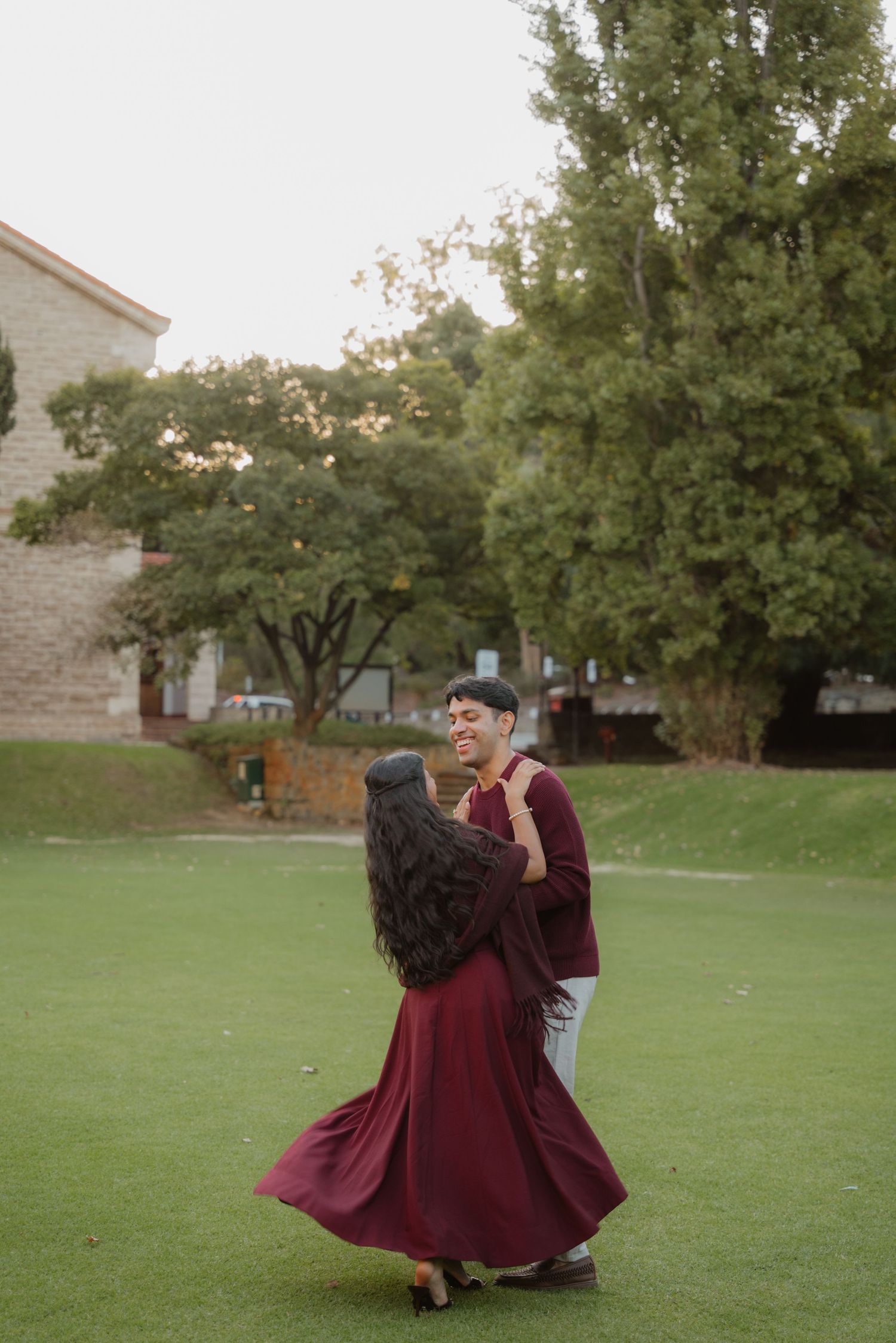 Joyful couple spinning on green lawn, woman in flowing burgundy dress, trees in background.