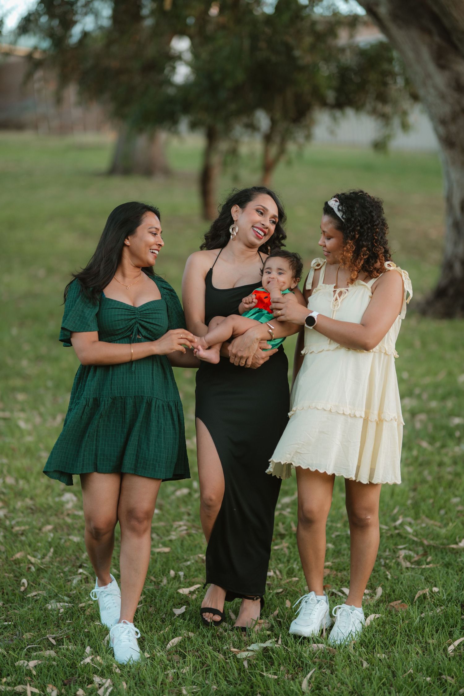Three smiling women in dresses standing outdoors in a park, one holding a baby with an orange flower.