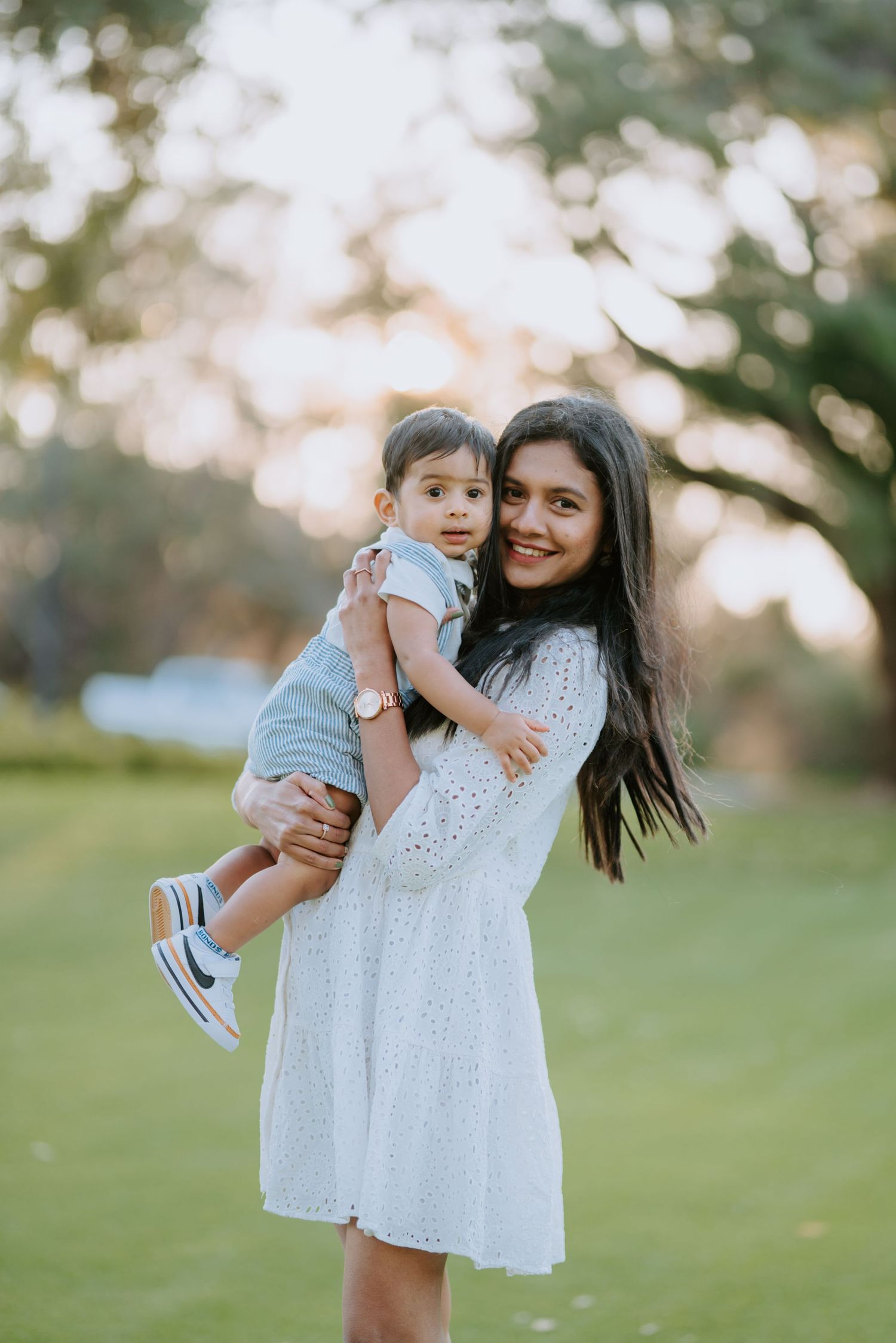 Smiling mother in white eyelet dress holds her baby boy at a sunny outdoor park.