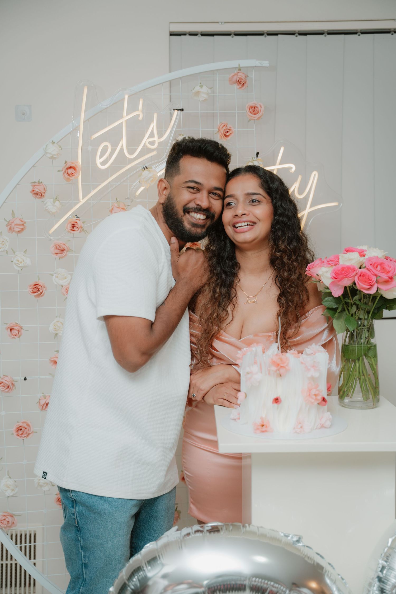 Happy couple posing together at a floral birthday party with neon Let's Party sign and pink roses.
