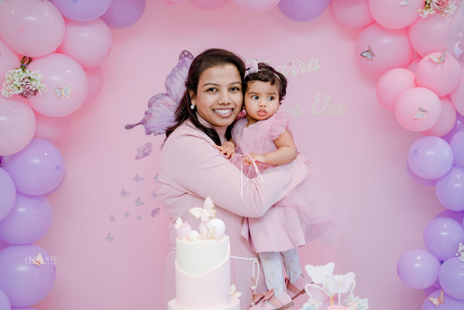 Smiling mother holding baby girl in matching pink outfits at butterfly-themed birthday party with balloons.