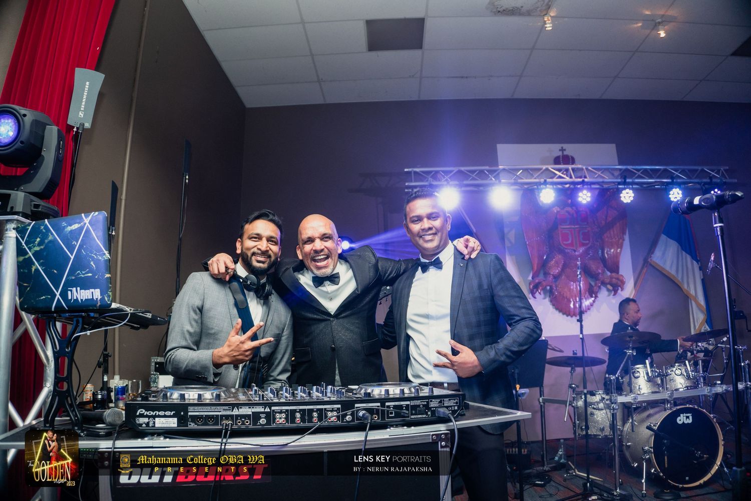 Three men smiling behind a DJ booth at an indoor evening event with stage lighting.