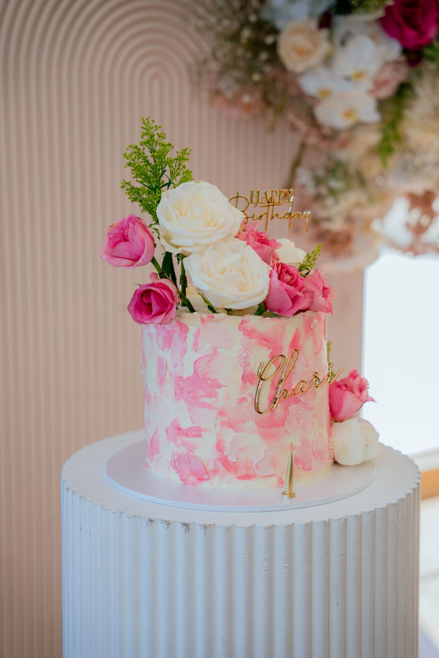 White birthday cake with pink watercolor design, gold name detail, and fresh pink and white roses on top.