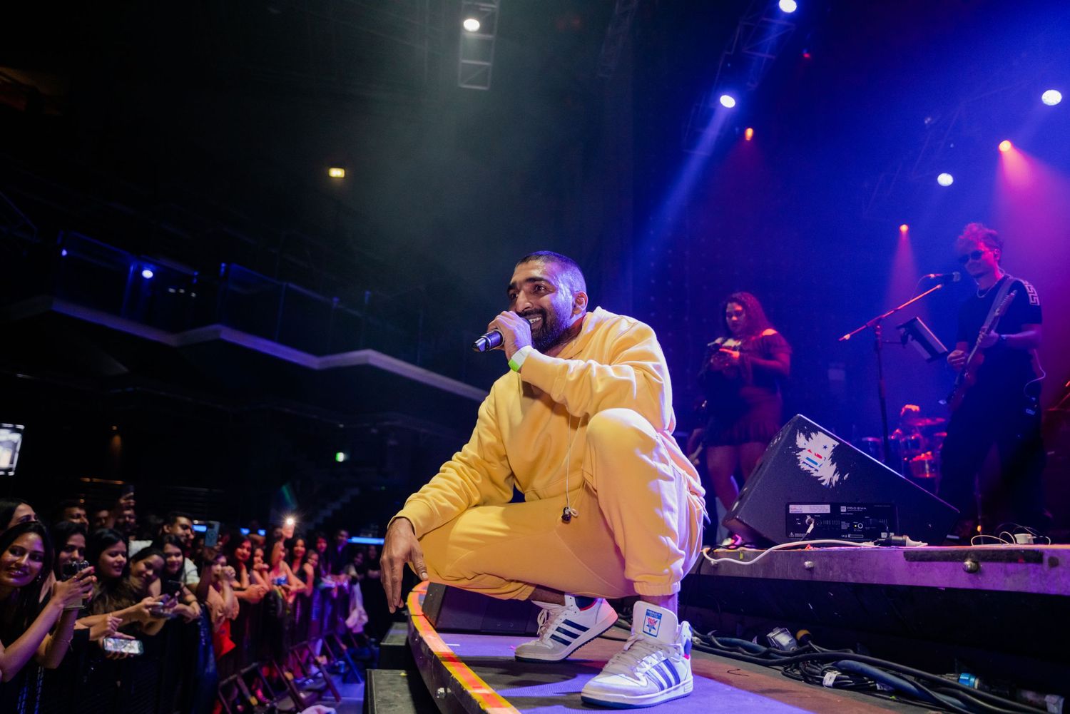 Artist in yellow Adidas tracksuit crouching on stage performing for crowd.