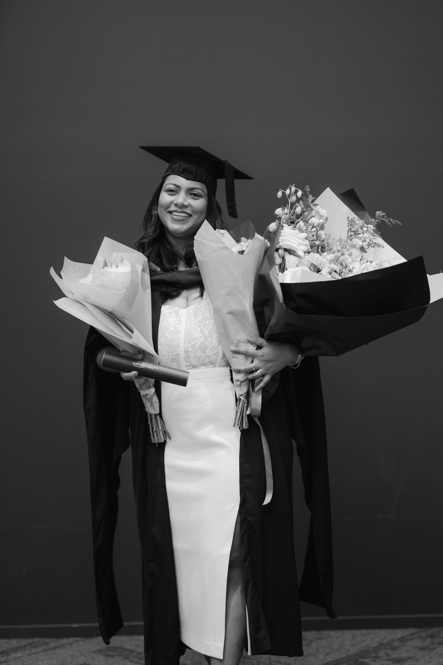 Black and white photo of a female graduate in cap and gown holding two bouquets of flowers.