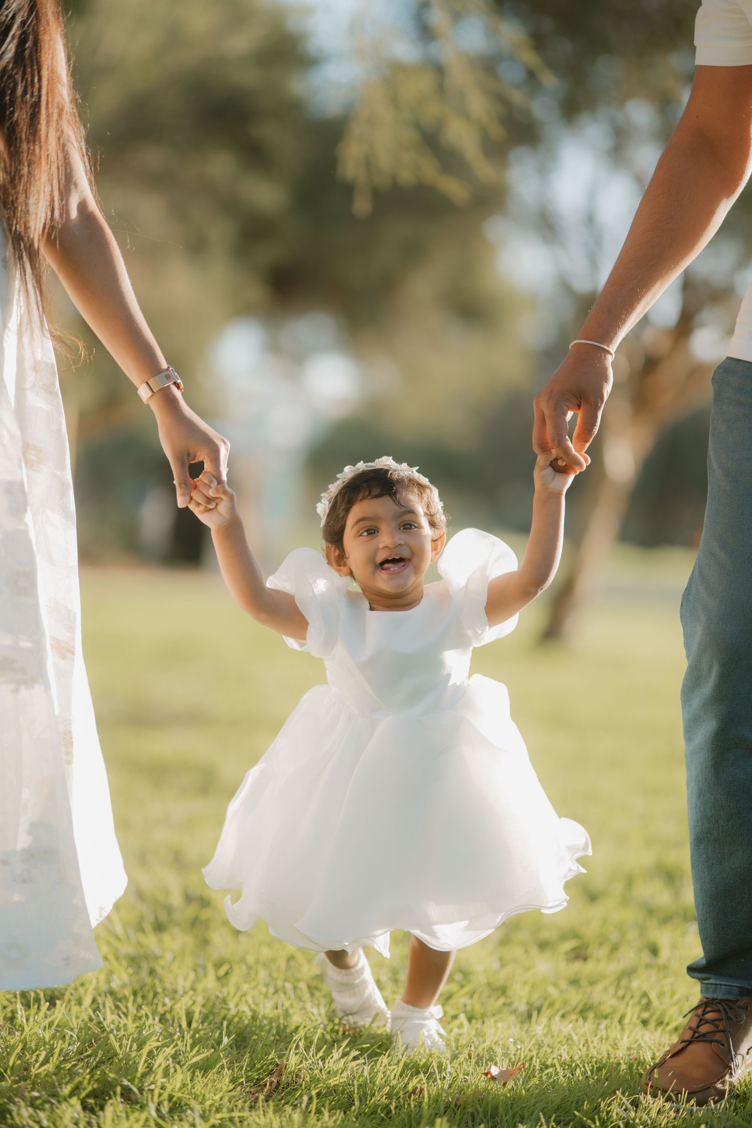 Toddler girl in white dress held by parents hands walking in sunny park.