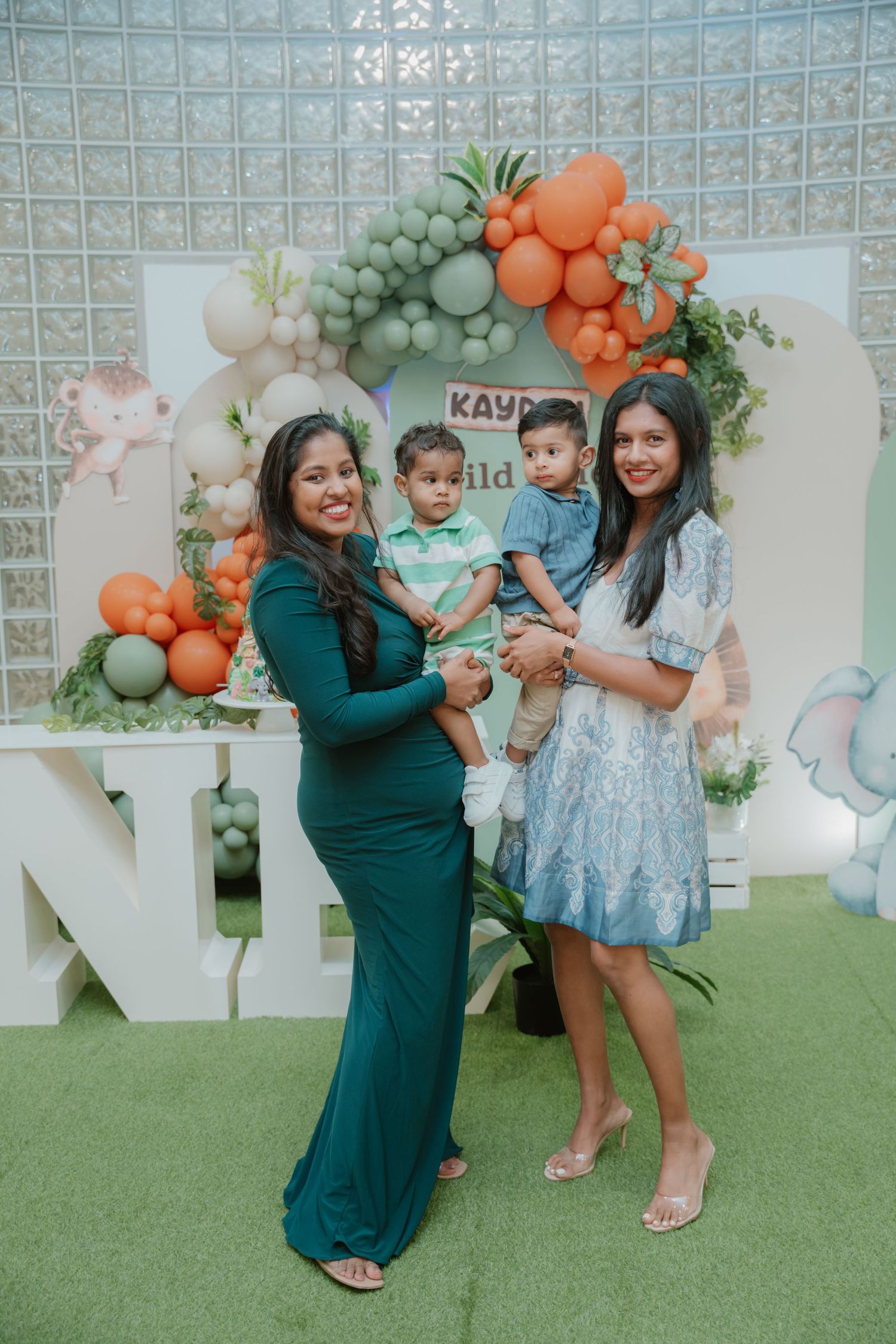 Two women each holding a baby boy pose at Kayden's Wild One safari-themed first birthday party backdrop.