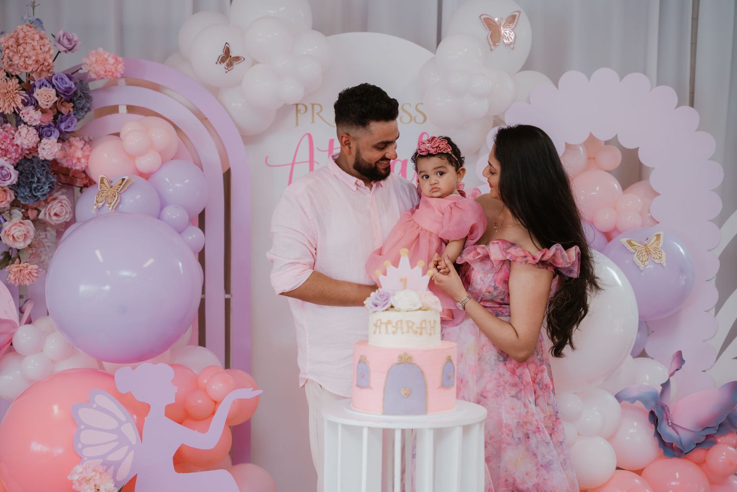 Parents with baby girl celebrating first birthday at pink princess themed party with cake.