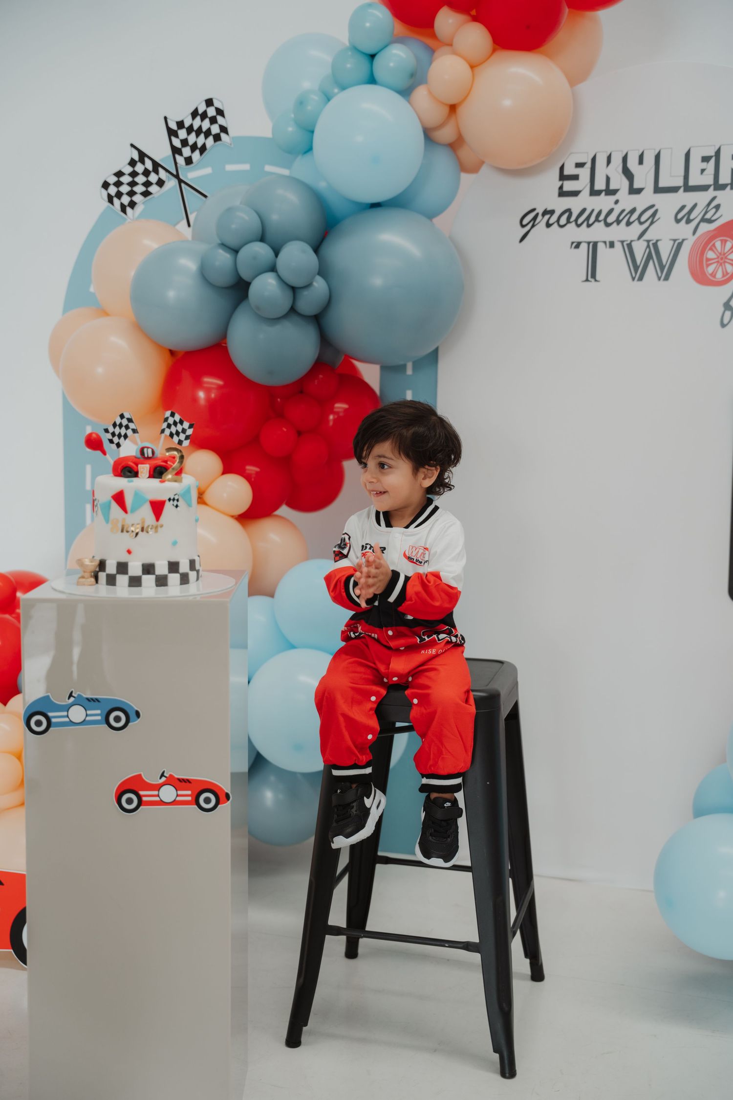 Toddler boy in racing outfit sits on stool at race car themed second birthday party with balloon arch.