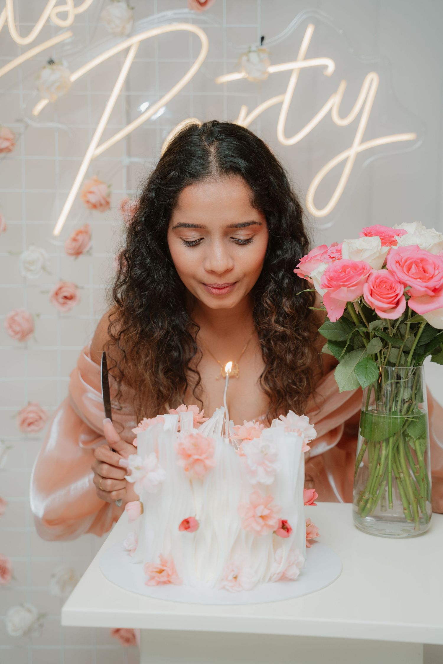Young woman blowing out candle on elegant white floral birthday cake at a Let's Party neon sign celebration.