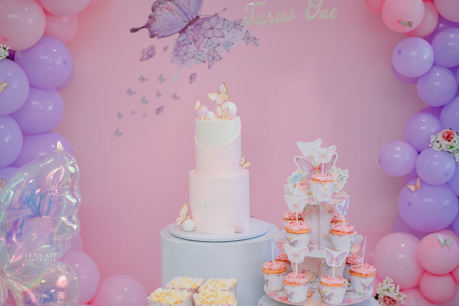 Elegant three-tier white butterfly birthday cake with cupcake tower and pink purple balloon arch backdrop.