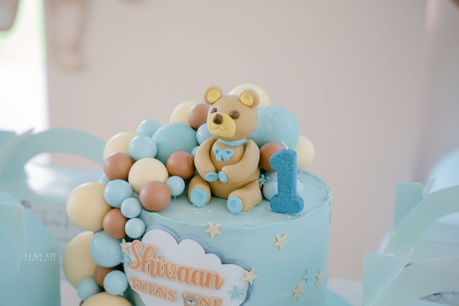 Close-up of a blue teddy bear first birthday cake with number one candle and colorful balloon details.
