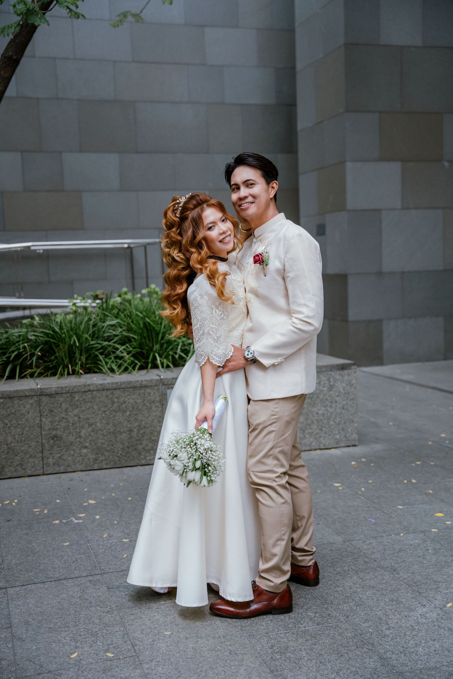 Smiling newlywed couple poses outdoors, bride in lace gown holding baby's breath bouquet.