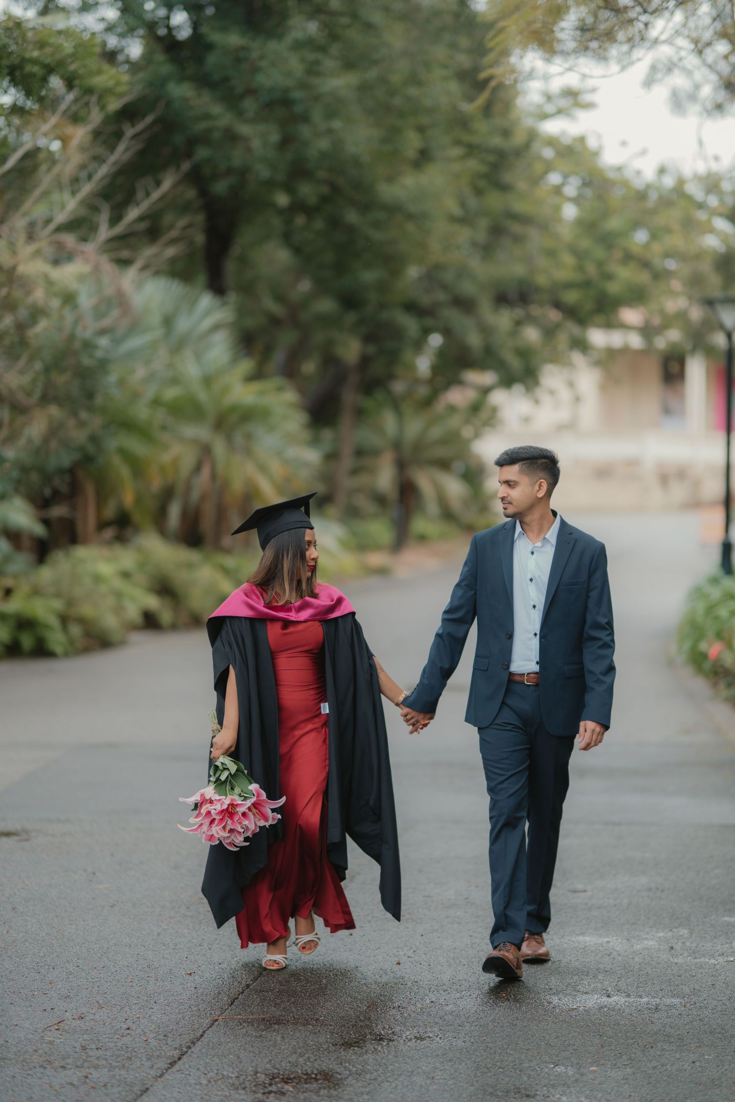 Graduate in black gown and red dress holding flowers walks hand-in-hand with suited partner outdoors.