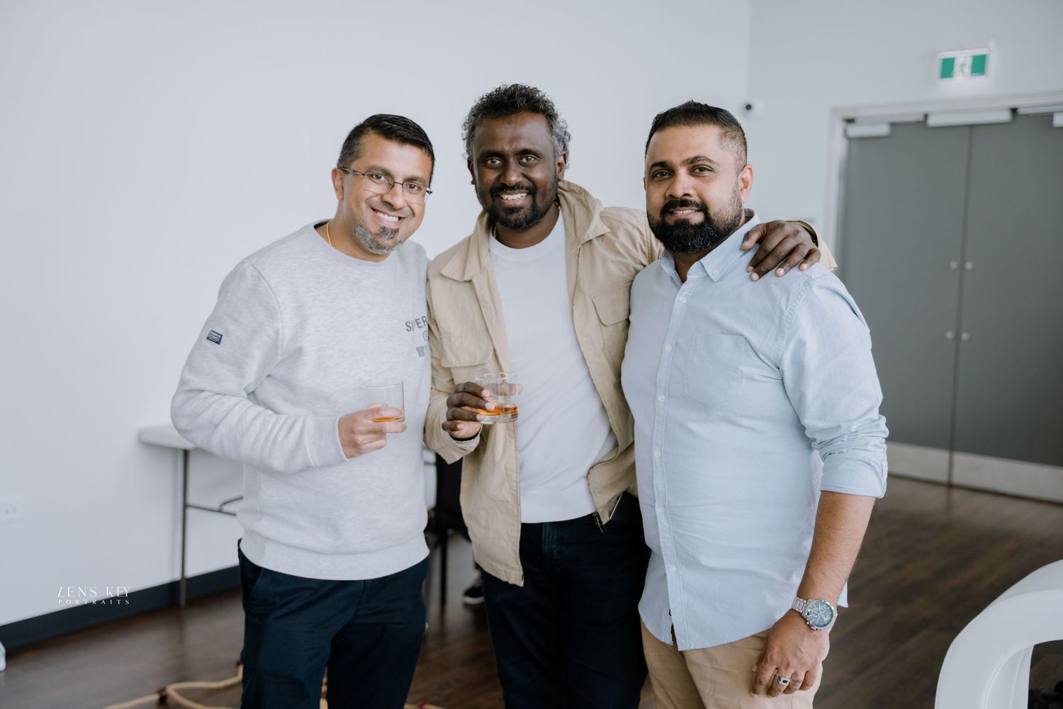 Three men smiling and posing together at an indoor celebration event with bright white background.